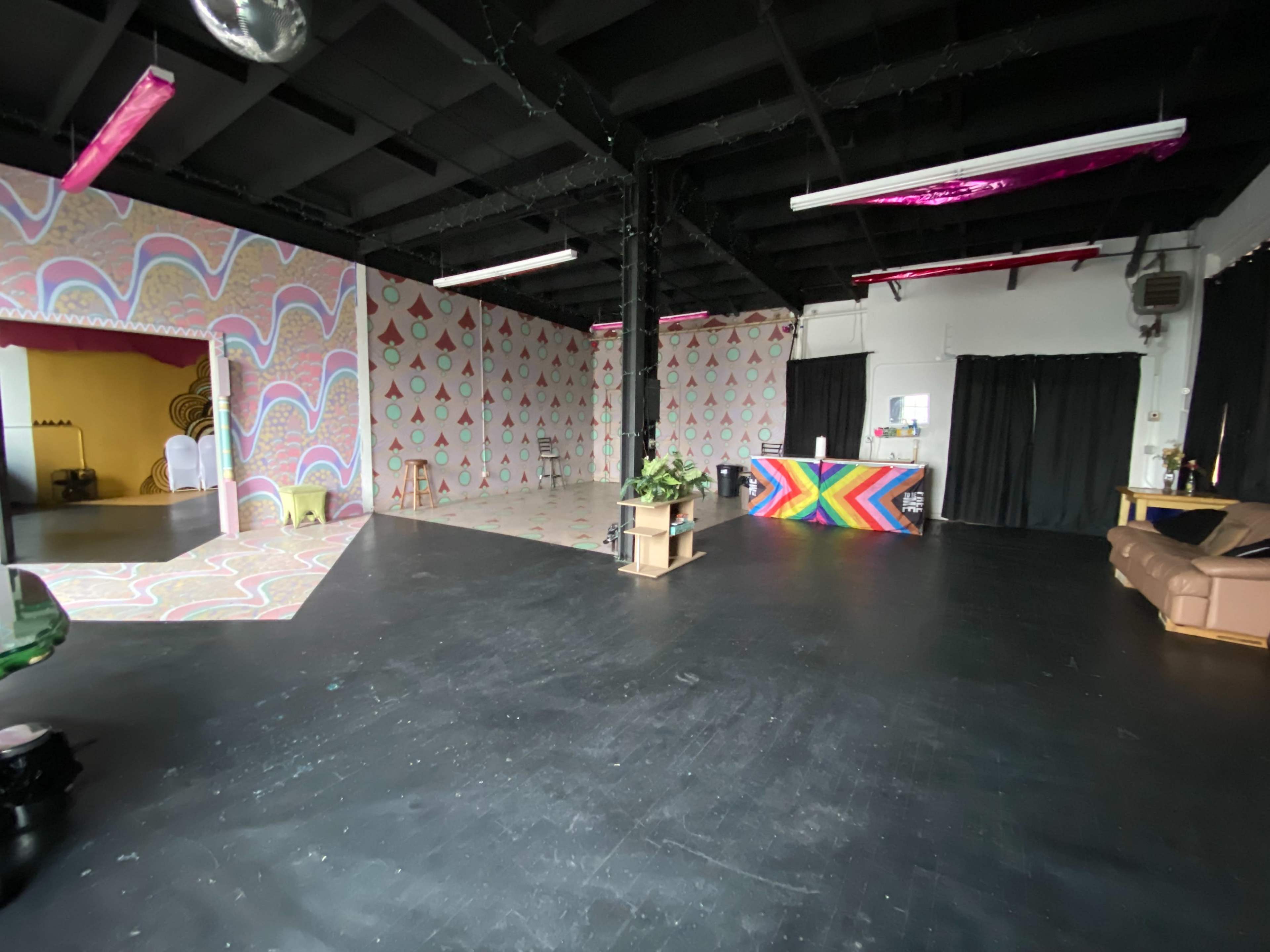 The image shows an empty studio space with colorful patterned walls, a black floor, and various furniture, including a couch and a reception area.