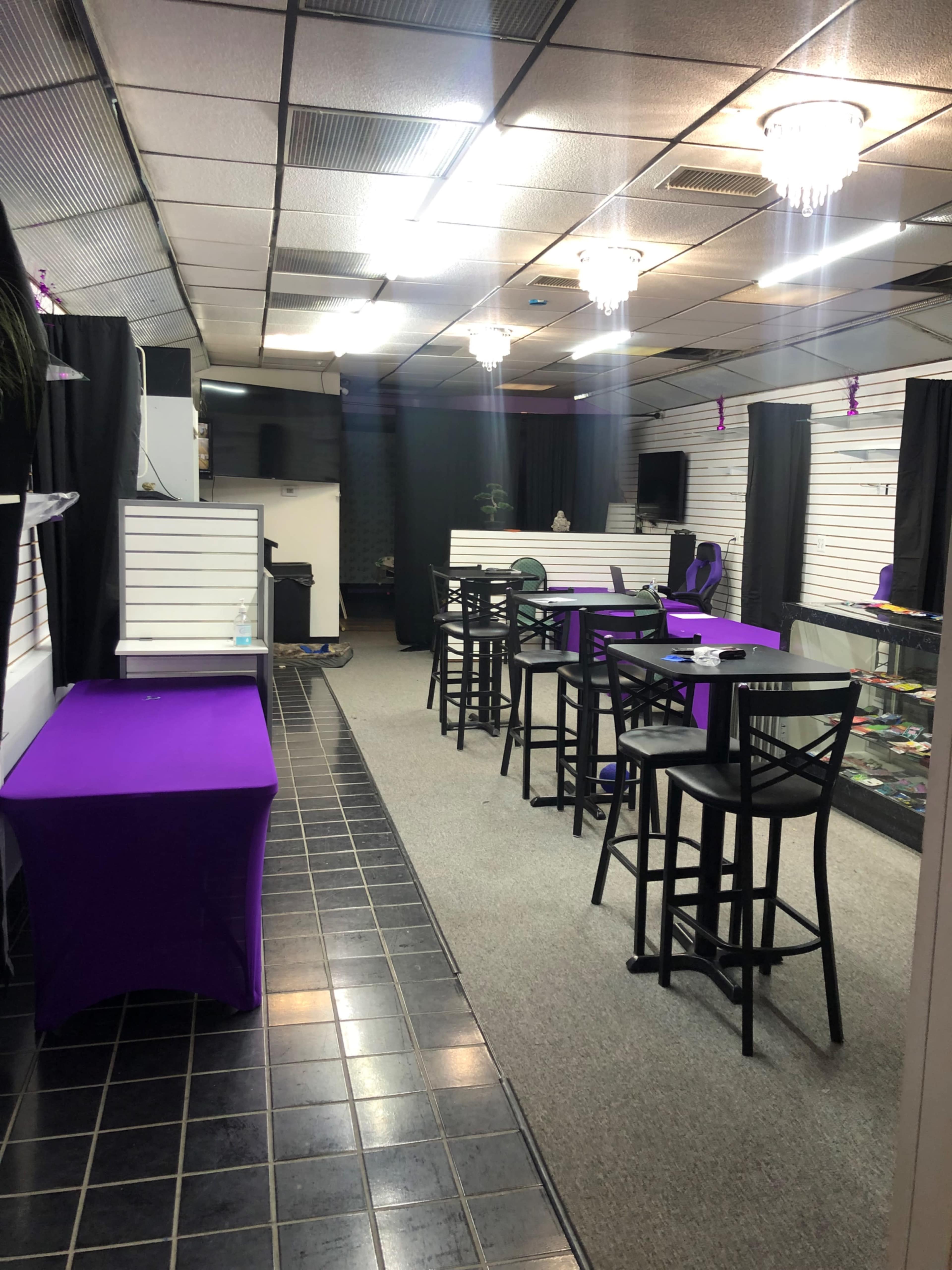 An empty retail space featuring black chairs and tables with purple tablecloths, displayed merchandise in glass cases, and illuminated by overhead lights.