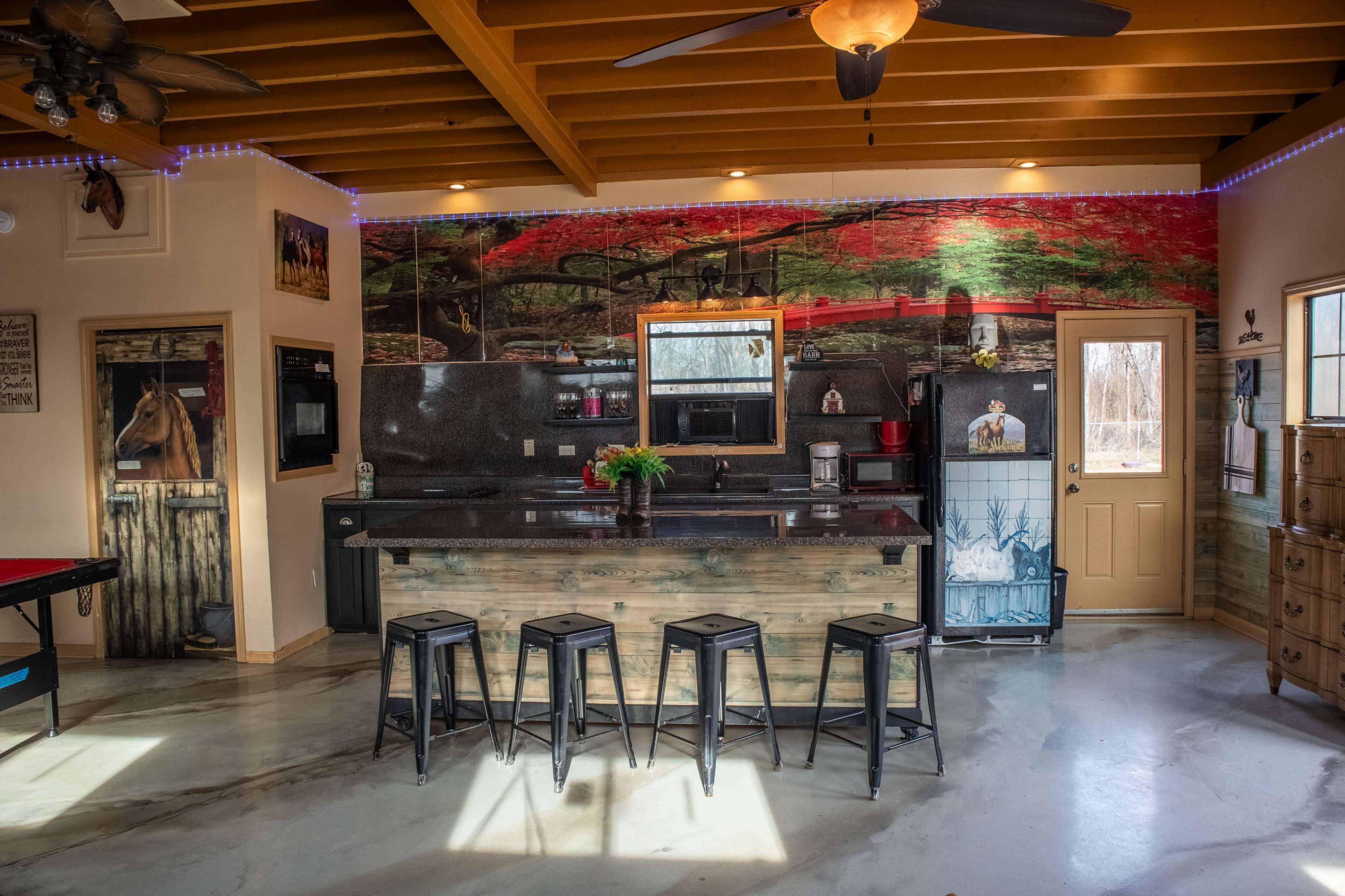 The image shows a spacious kitchen area featuring a bar-style countertop with several stools, modern appliances, and a large mural depicting a scenic landscape on the wall.