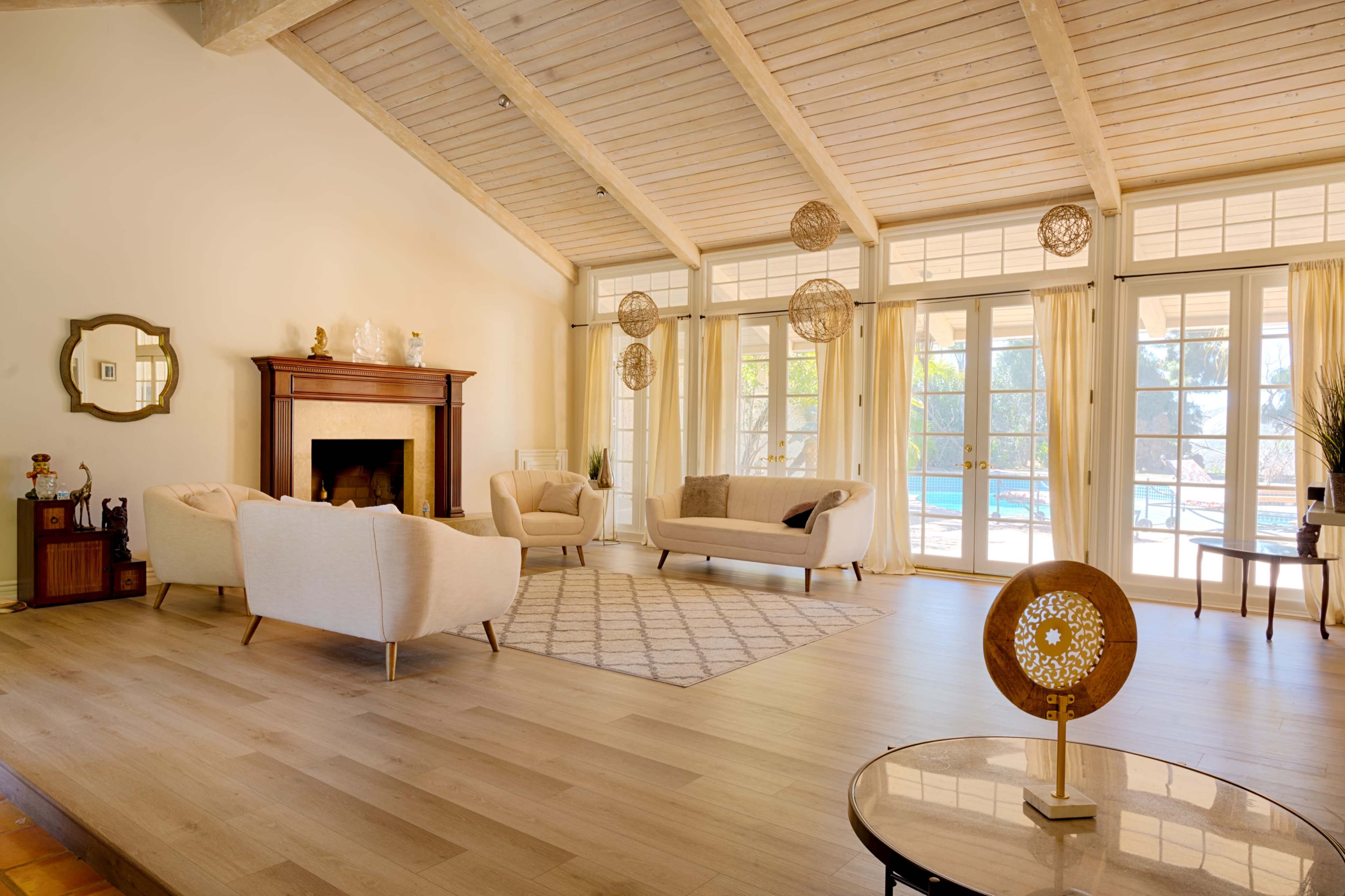 A spacious living room features a fireplace, two sofas, a patterned area rug, and large windows overlooking a pool area.