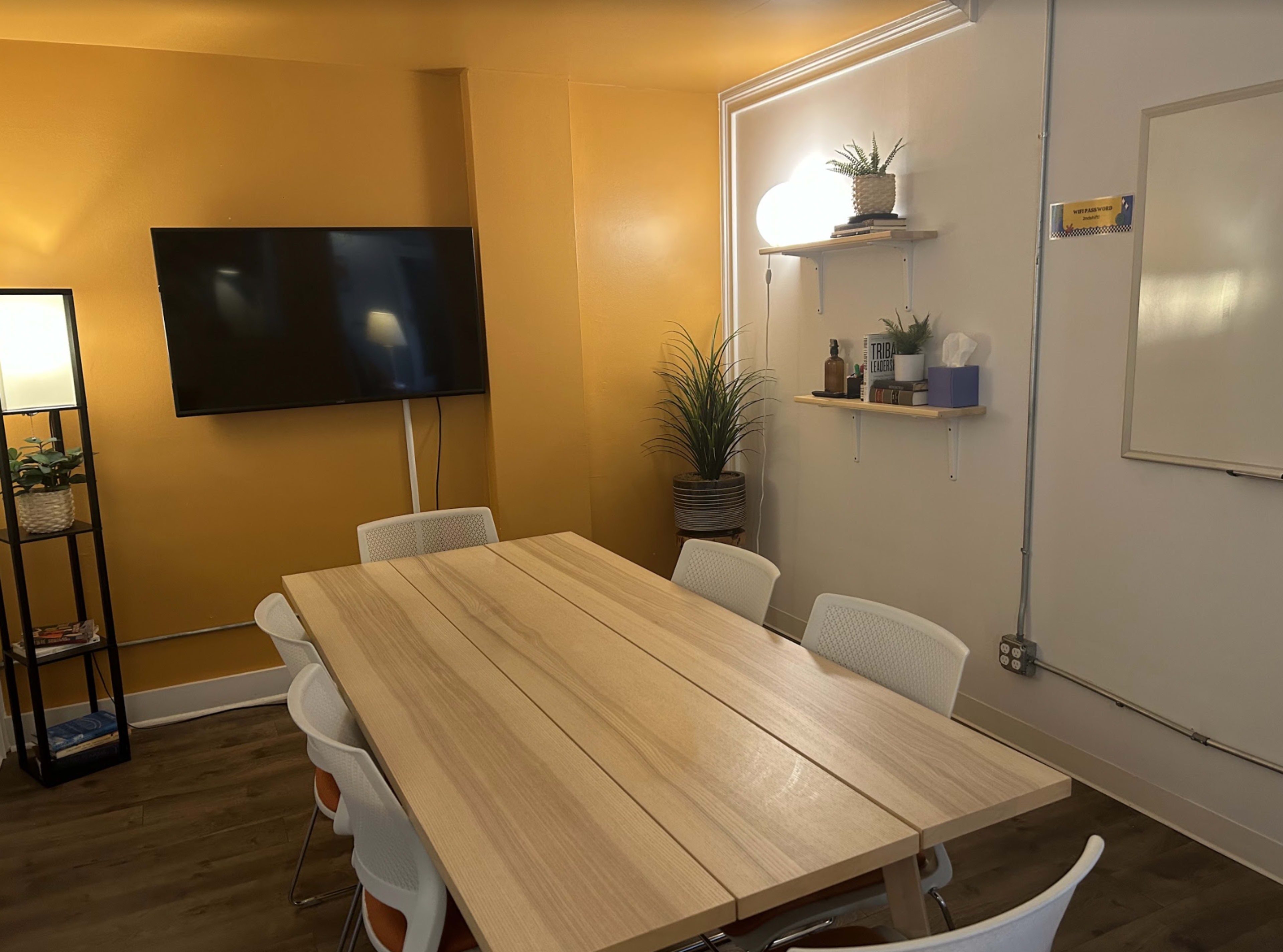 A conference room with a long wooden table, white chairs, a wall-mounted TV, and shelves with plants and decor.