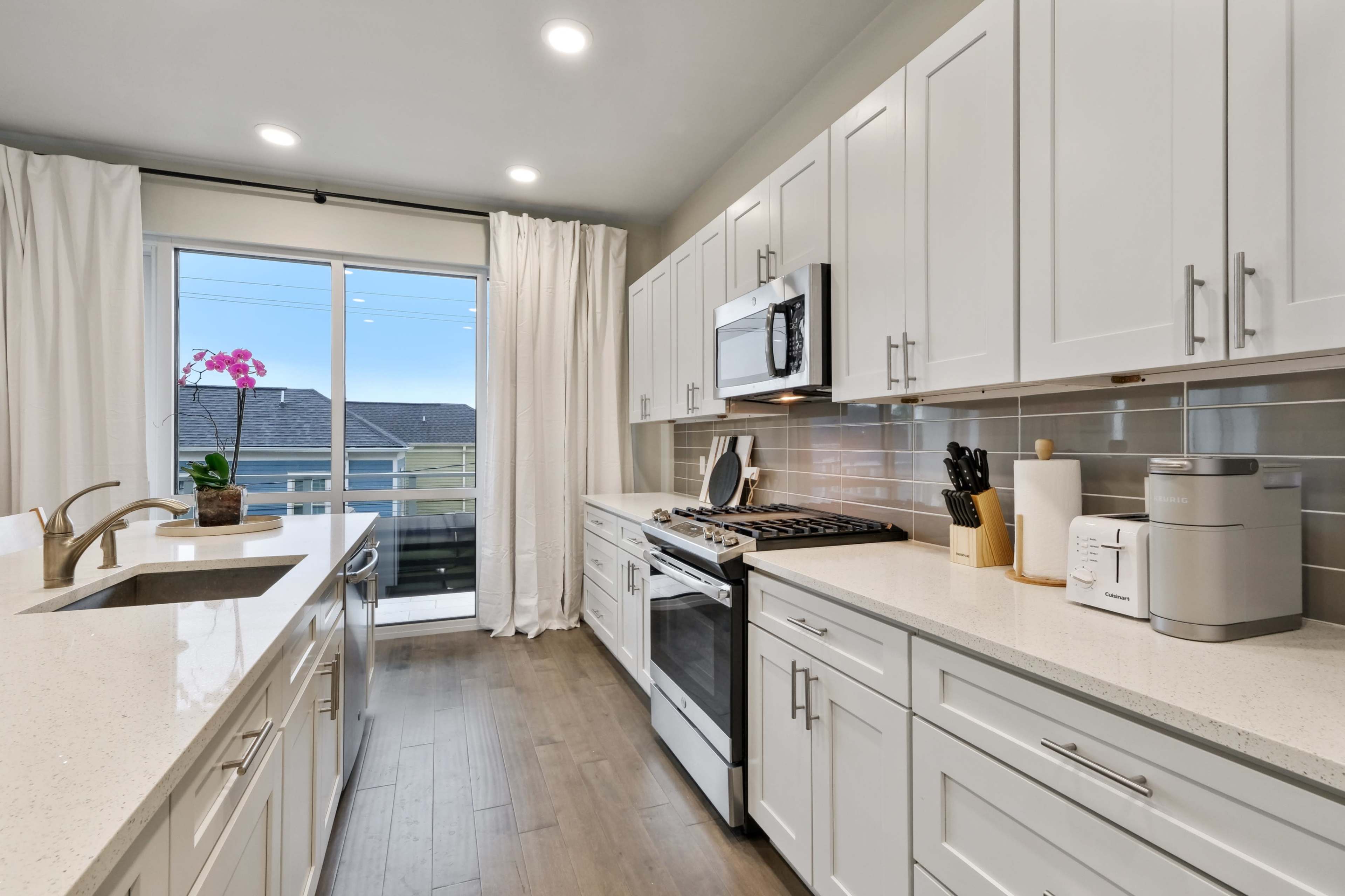 A modern kitchen features white cabinetry, stainless steel appliances, and a large window with sheer curtains.