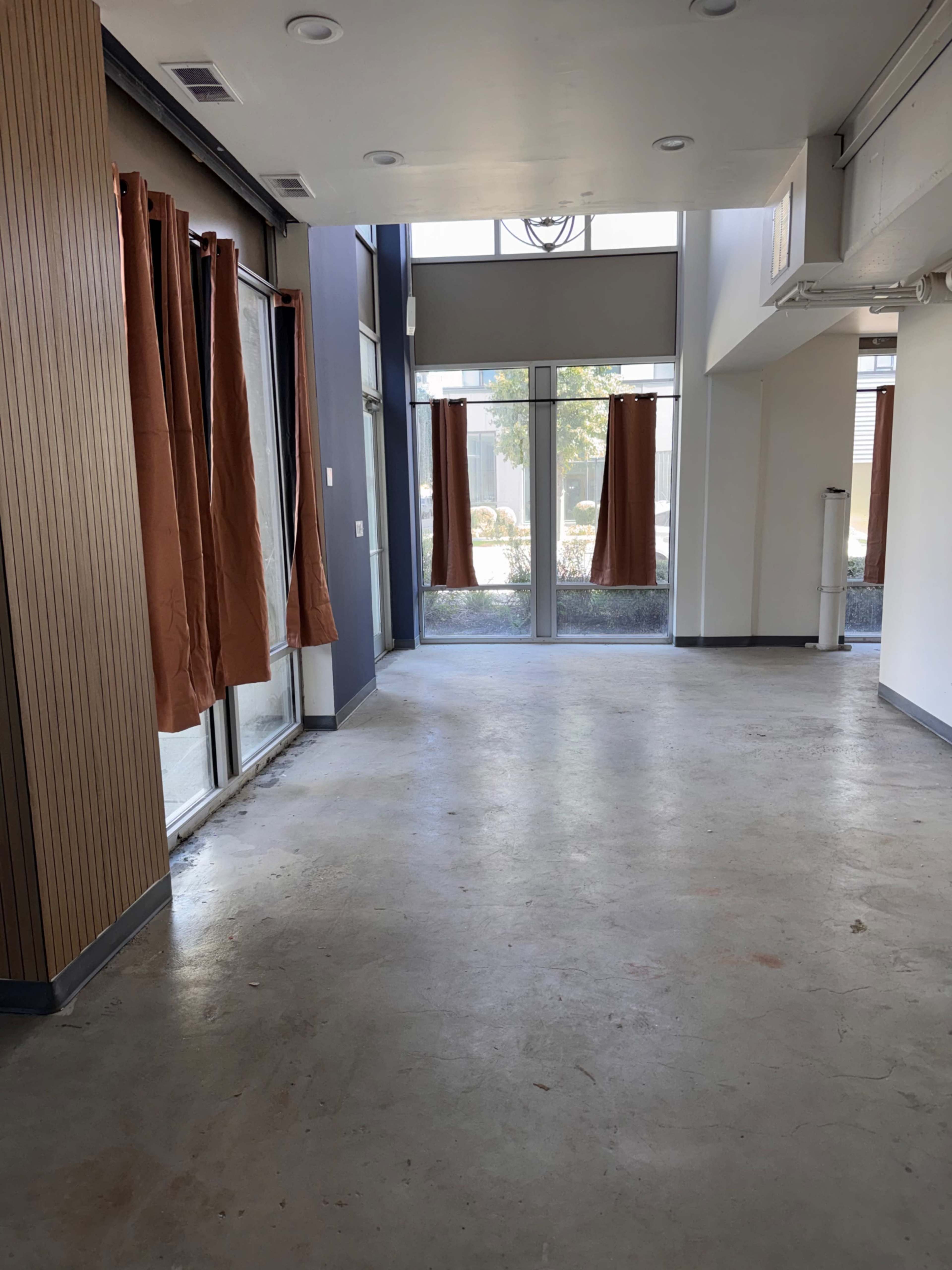 The image shows an empty interior space with concrete flooring, large windows covered by brown curtains, and minimal furnishings.