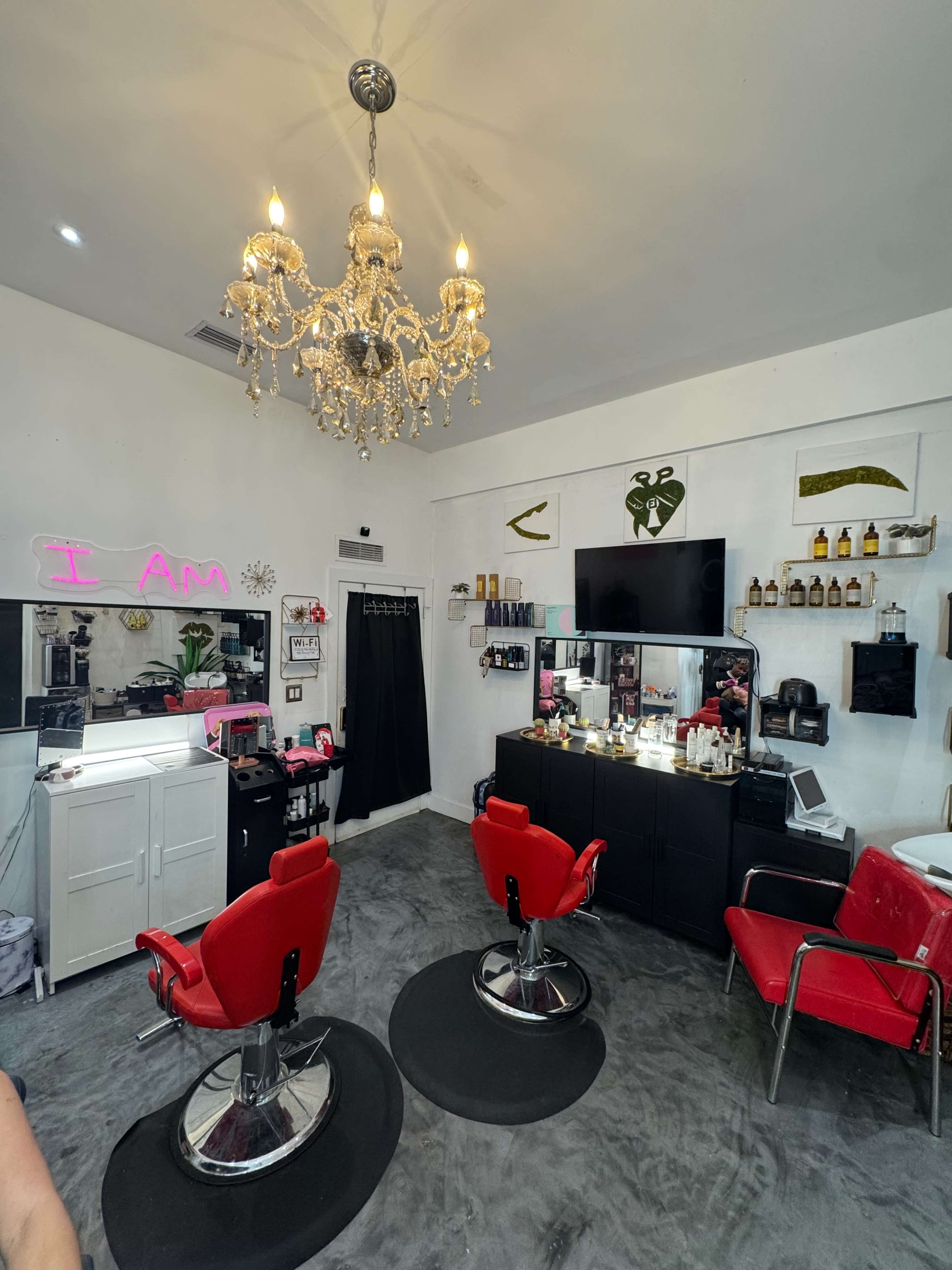 The image shows a brightly lit hair salon featuring two red styling chairs, a chandelier, and various beauty products displayed on shelves.
