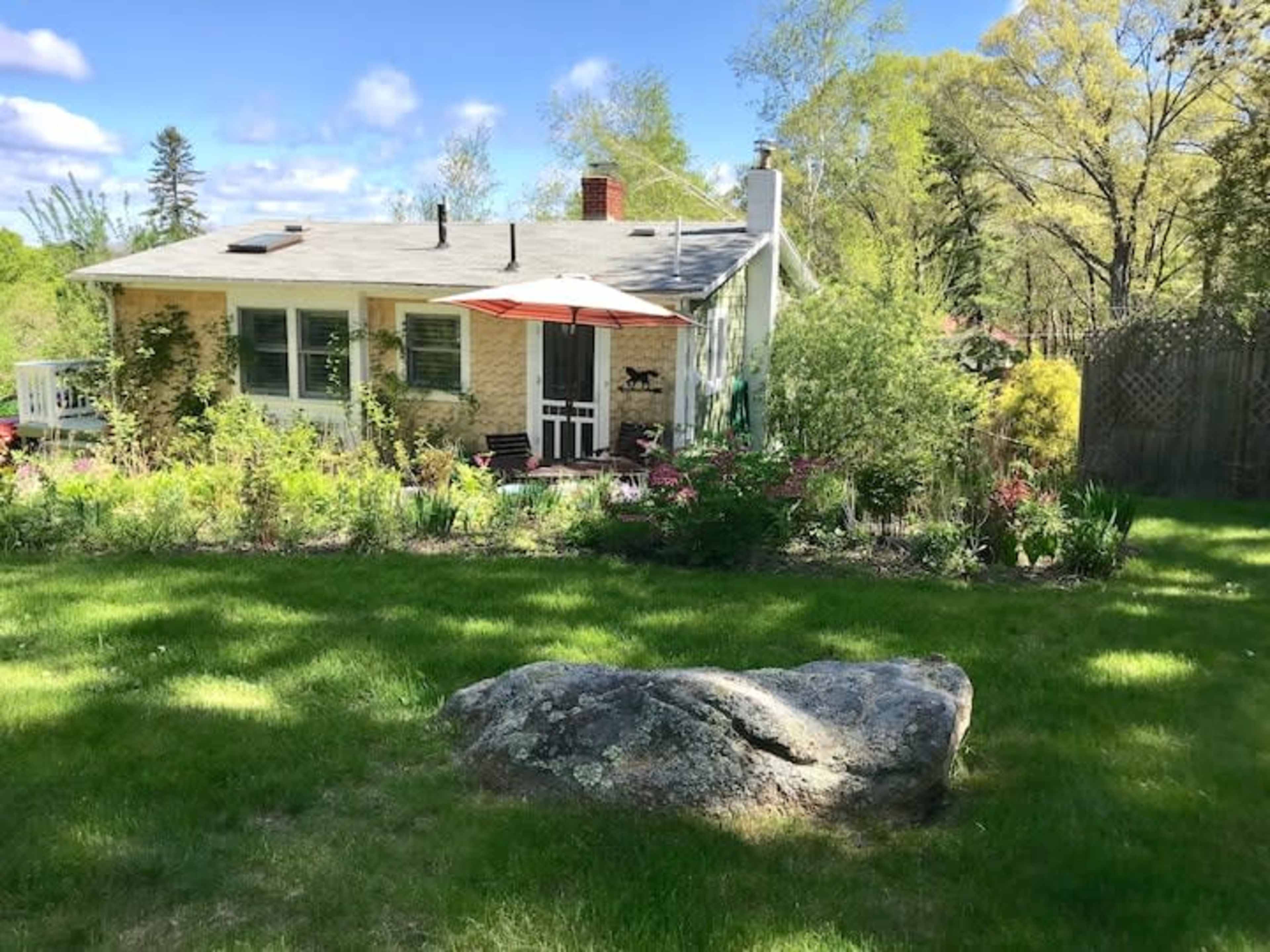 A single-story house with a gray roof and an orange umbrella stands in a garden surrounded by various plants and a large rock in the foreground.