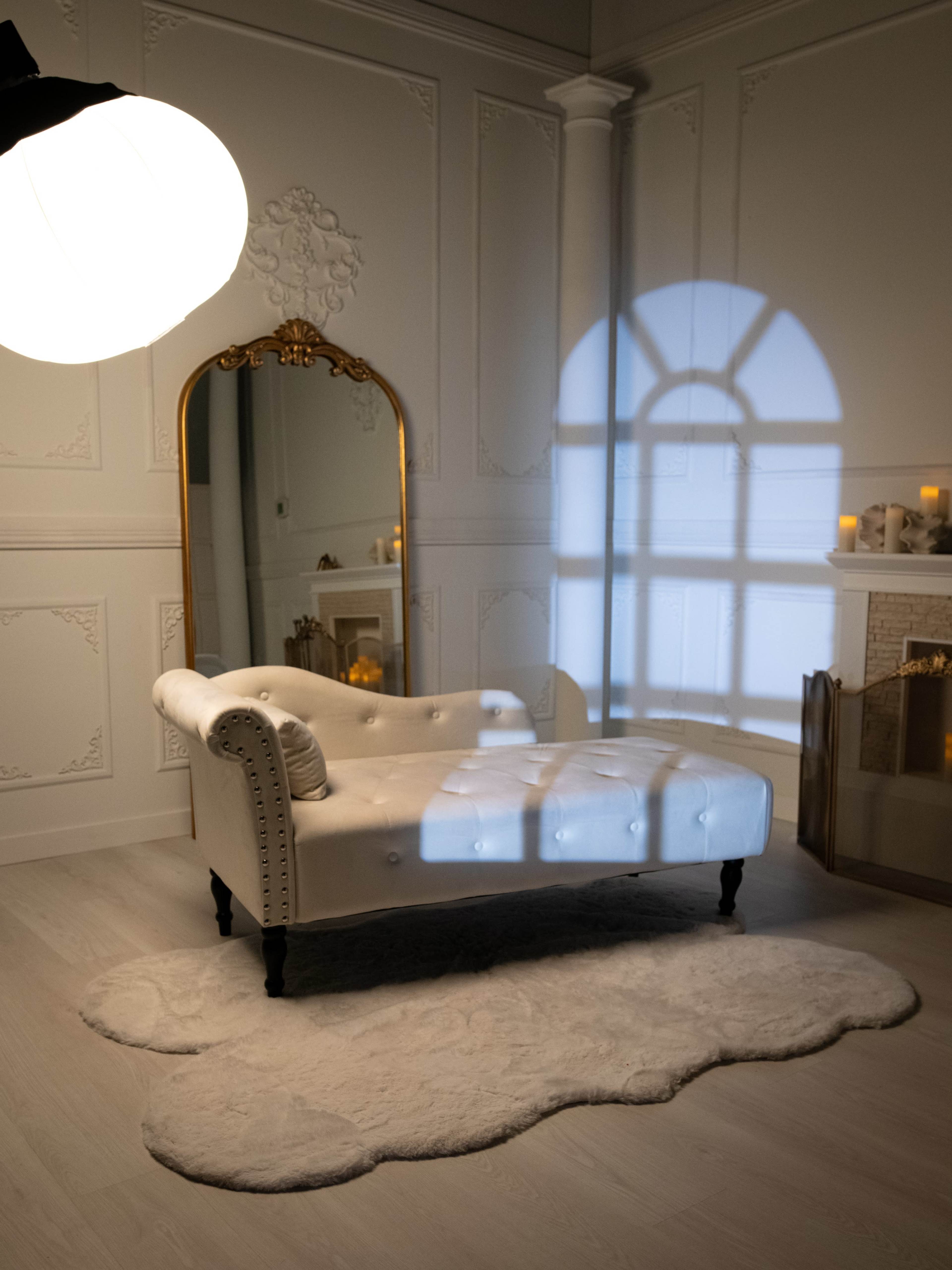 The image features a white chaise lounge in a softly lit room with a large mirror and a shadow cast by a window on the wall.