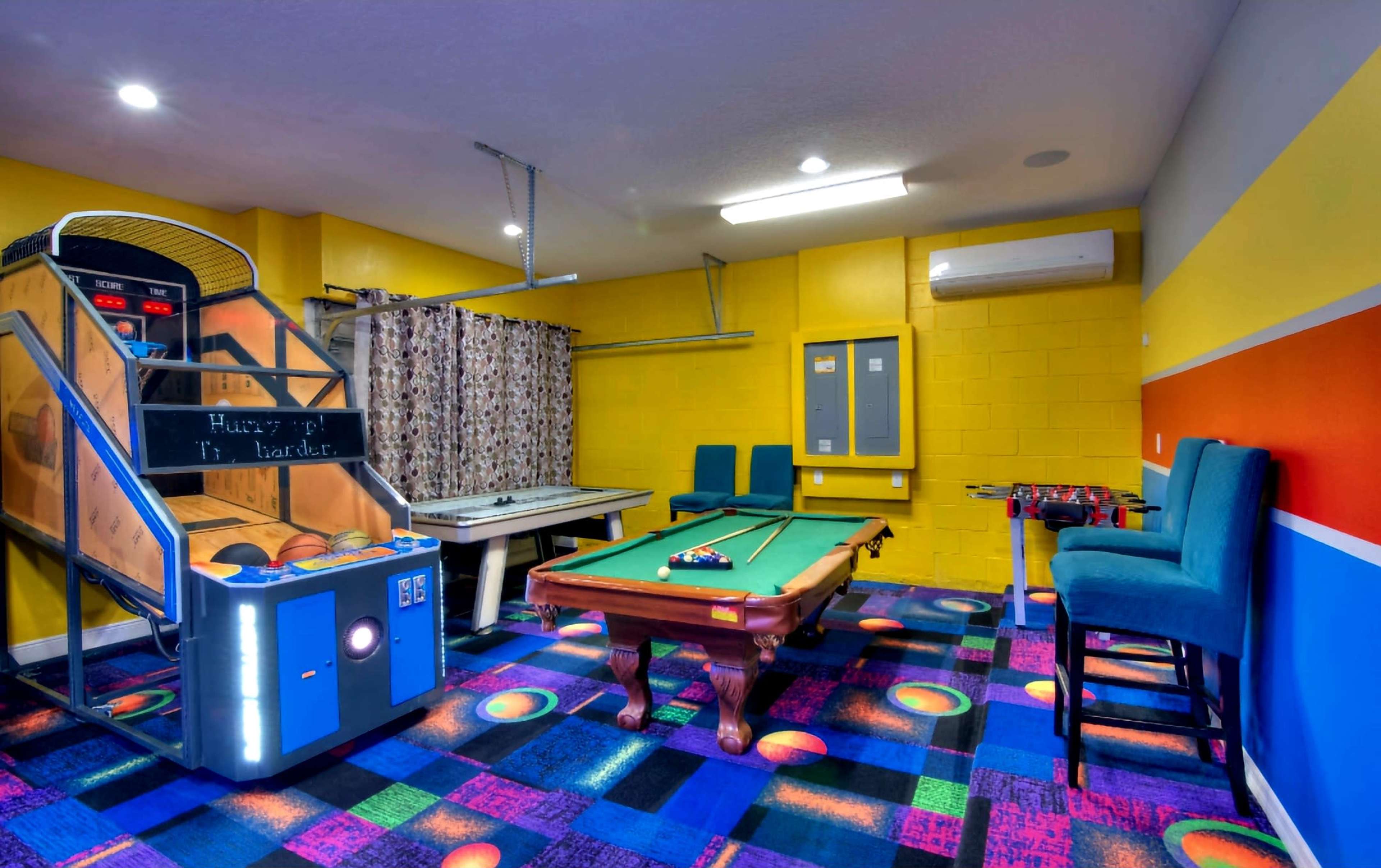 The image shows a recreational room featuring an arcade basketball game, a pool table, and colorful carpet tiles.