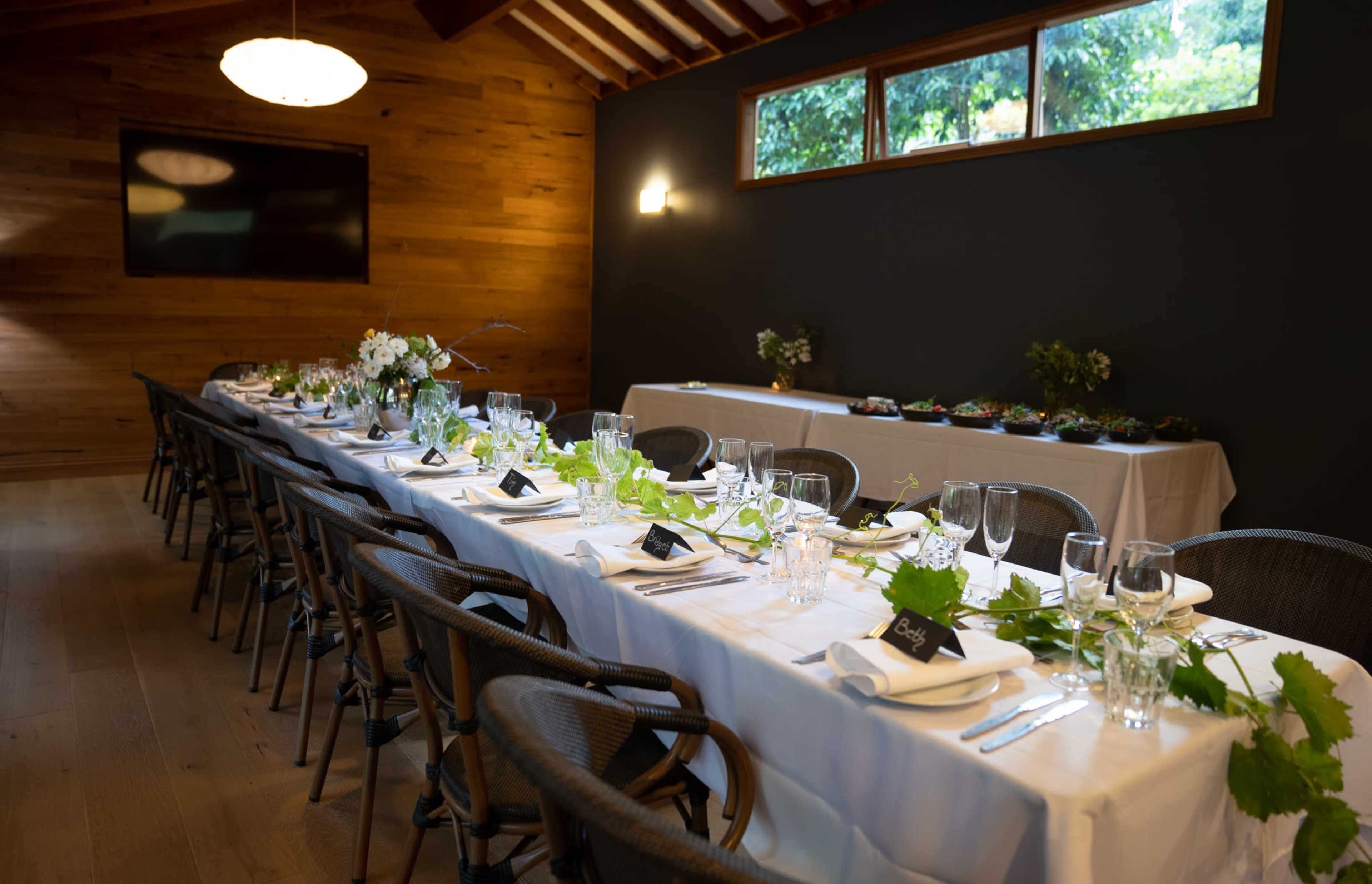 A long dining table is set with white tablecloths, glassware, and floral arrangements in a wooden room with a dark wall and a large screen.