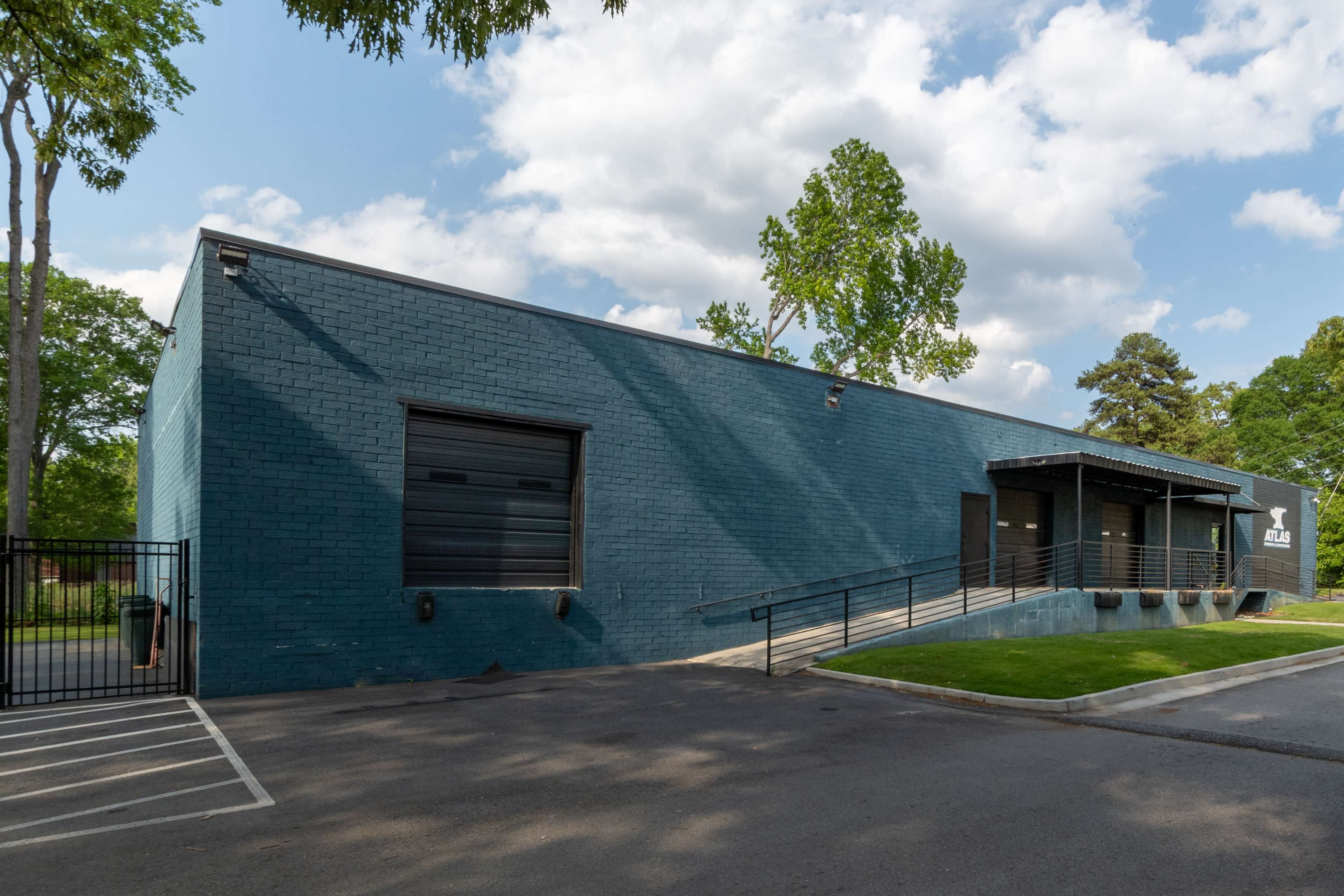 A large, blue brick building with three loading dock doors and a sloped ramp is situated beside a paved parking area and surrounded by trees.