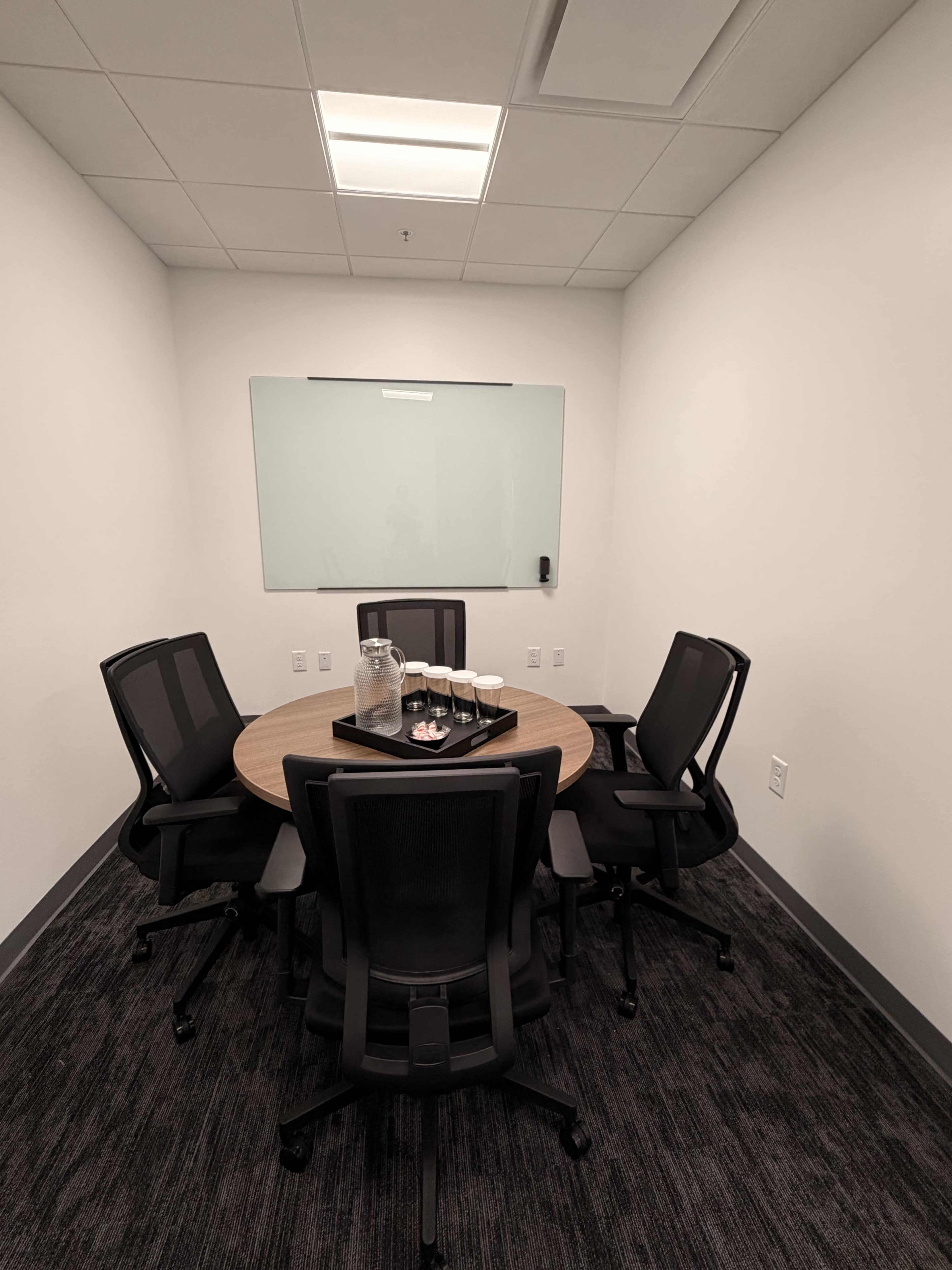 A small conference room features a round wooden table surrounded by five black office chairs, with a glass pitcher and cups on a tray in the center.