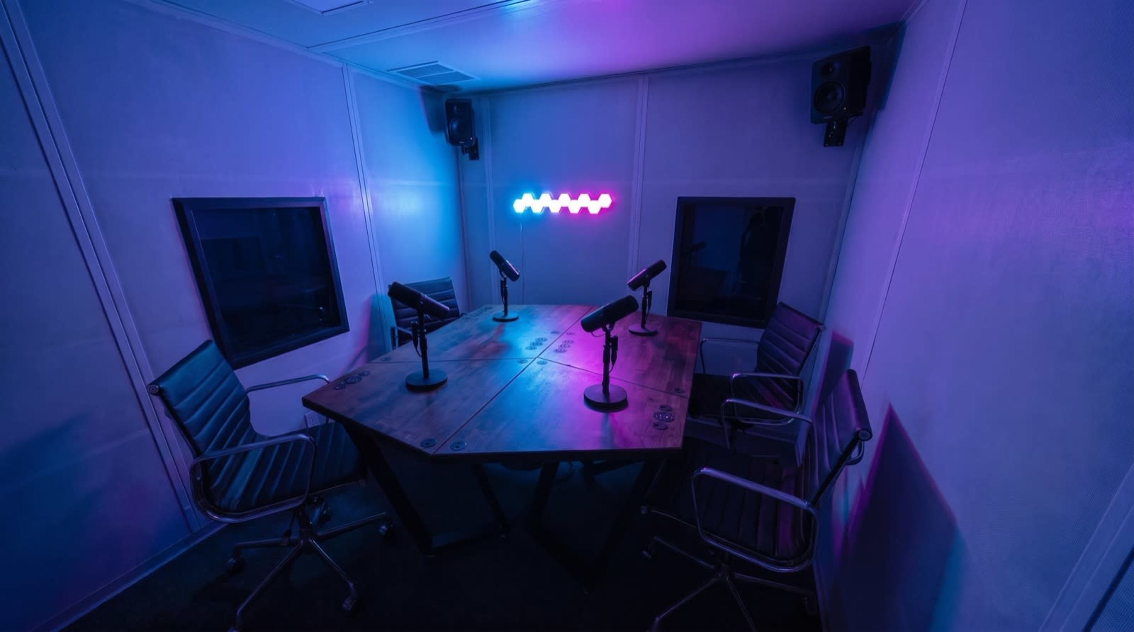 The image shows a modern audio recording studio with a large wooden table surrounded by six chairs and equipped with several microphones, illuminated by colorful lights.