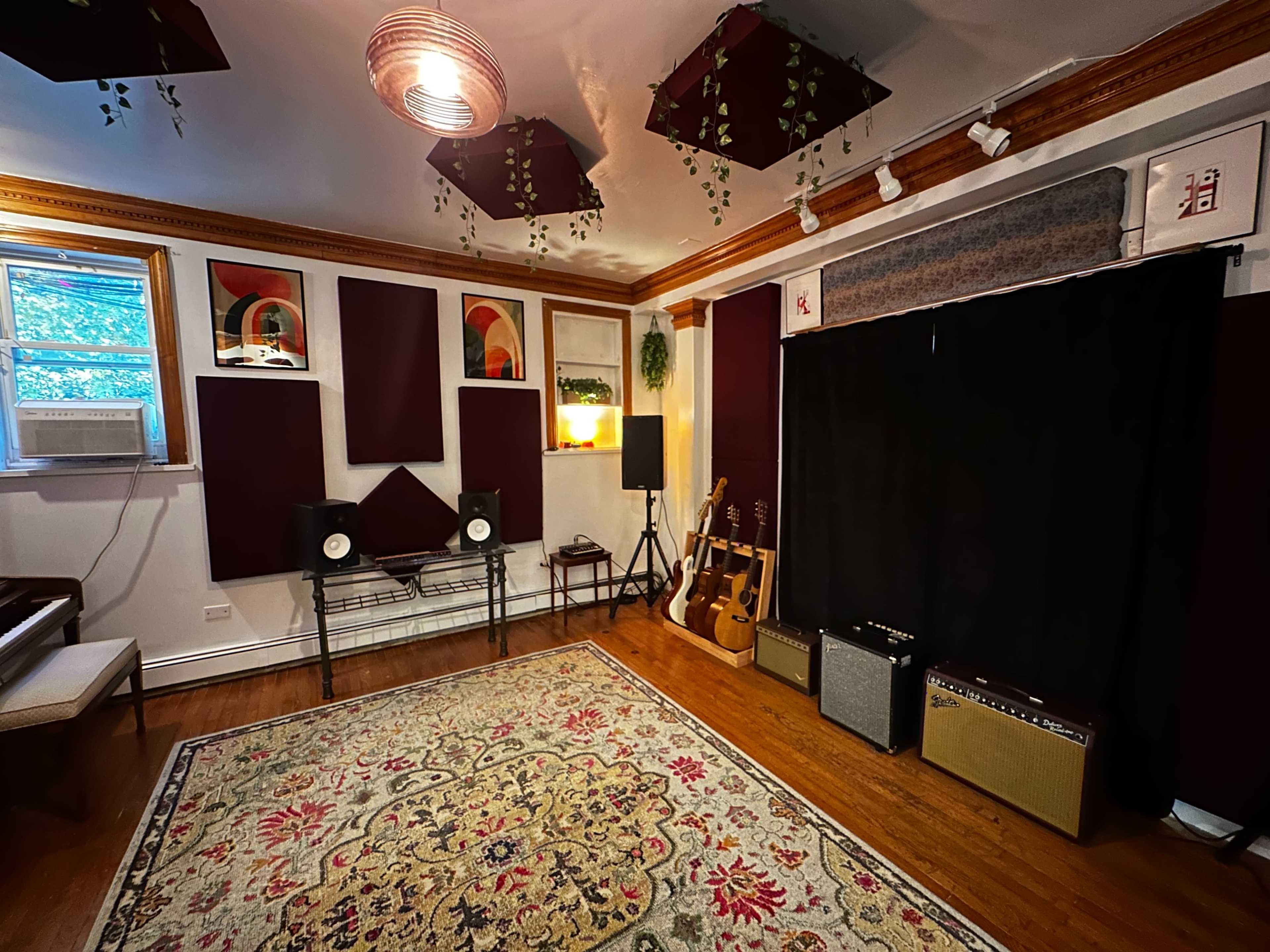 The image shows a cozy music studio with a patterned rug, instruments, speakers, and soundproofing panels on the walls.