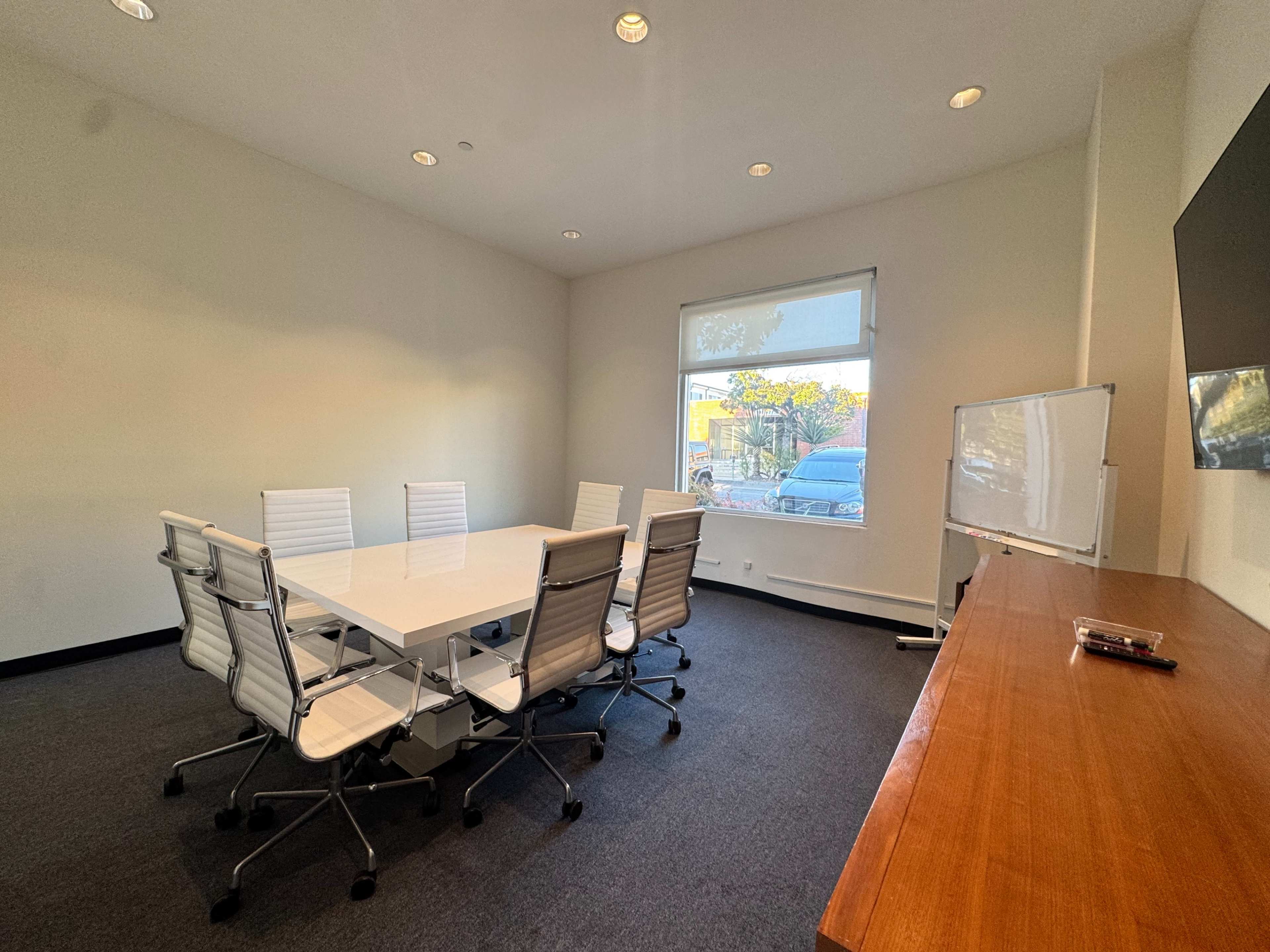 The image shows a modern conference room featuring a large white table surrounded by eight white chairs, a window with natural light, and a wooden cabinet with a television mounted on the wall.