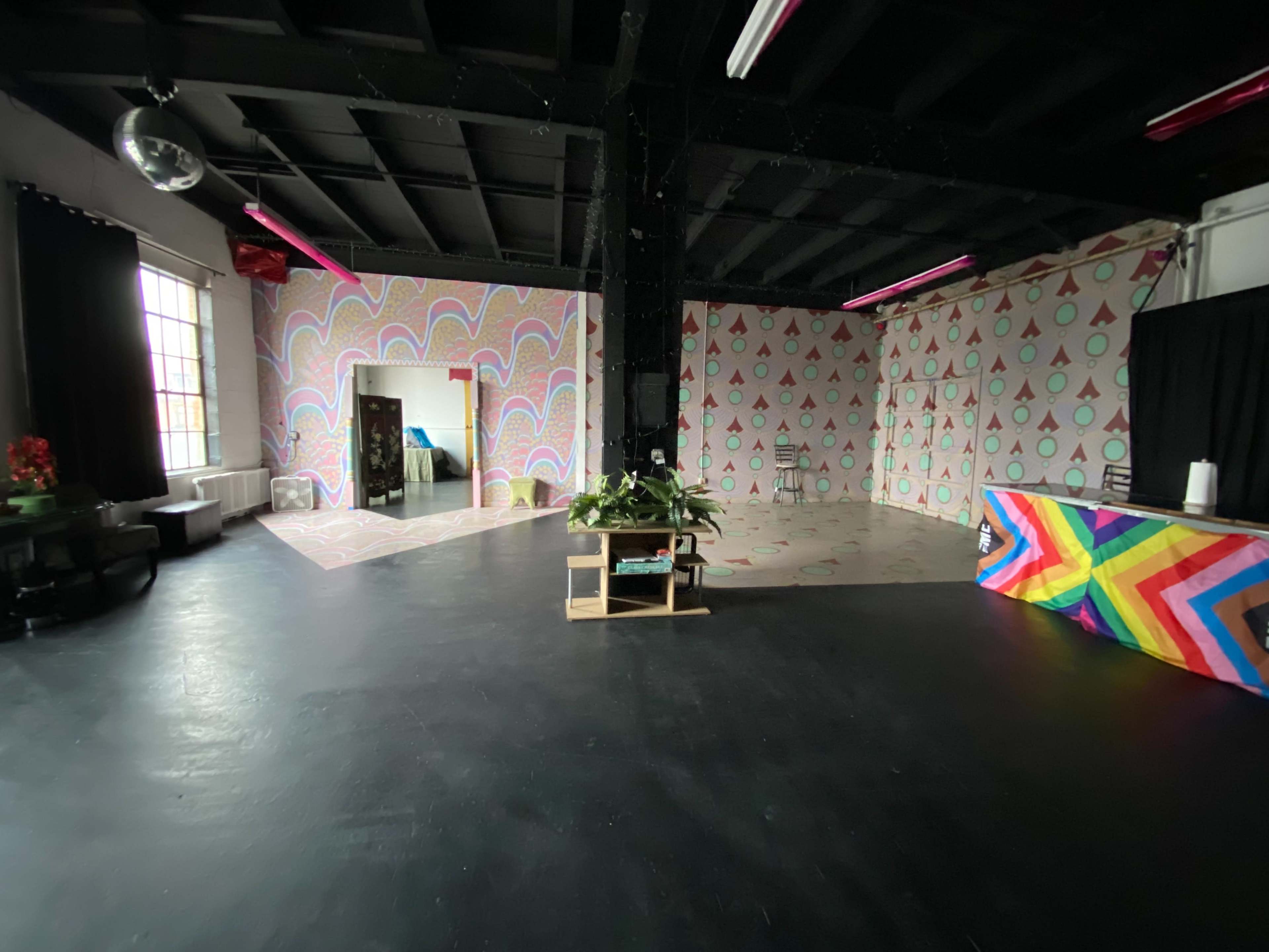The image shows a spacious room with colorful patterned walls, a dark floor, and a small stage area with a vibrant rainbow backdrop.