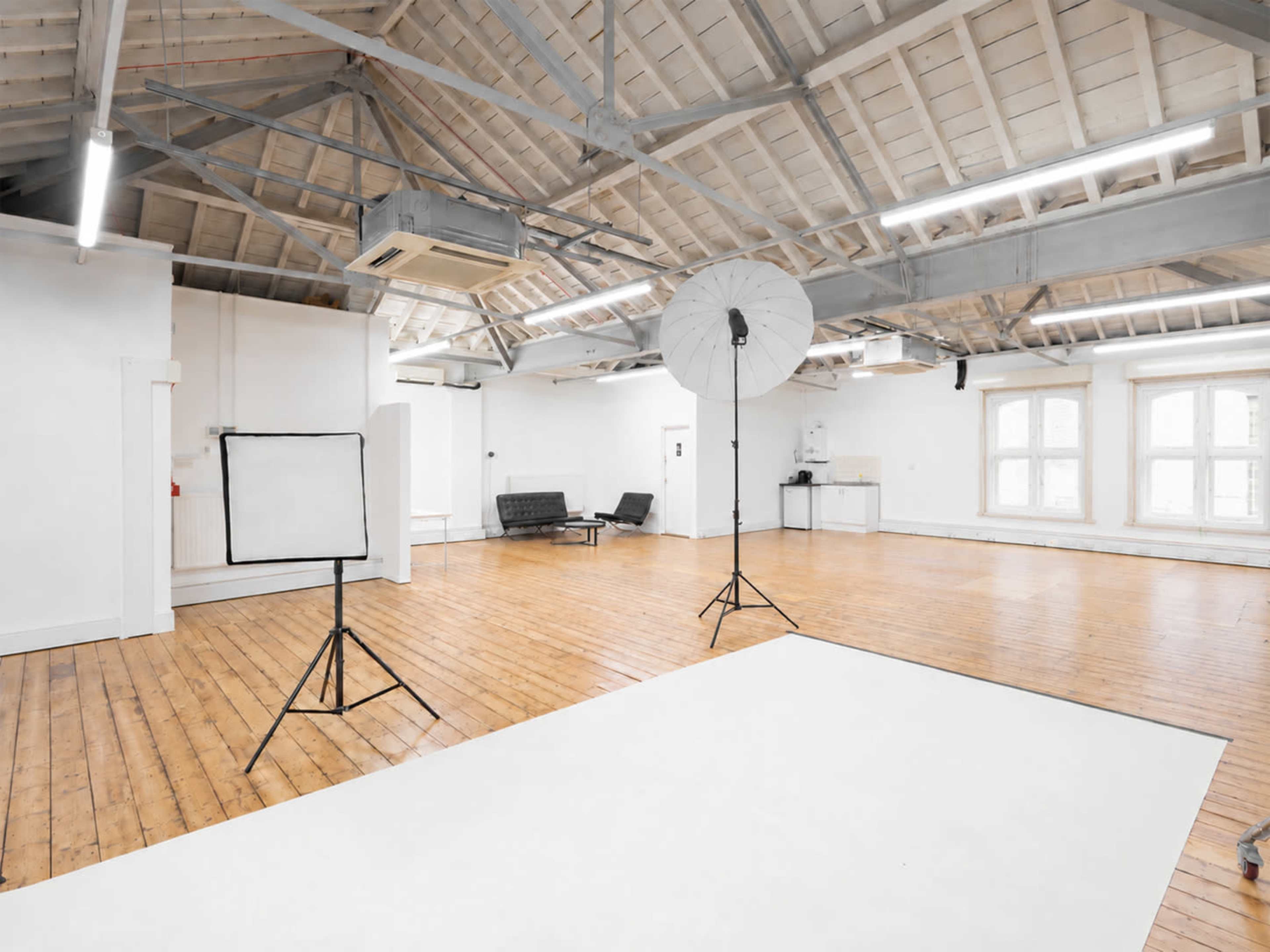 The image shows a spacious, well-lit photography studio with wooden floors, a white backdrop, and two lighting setups.