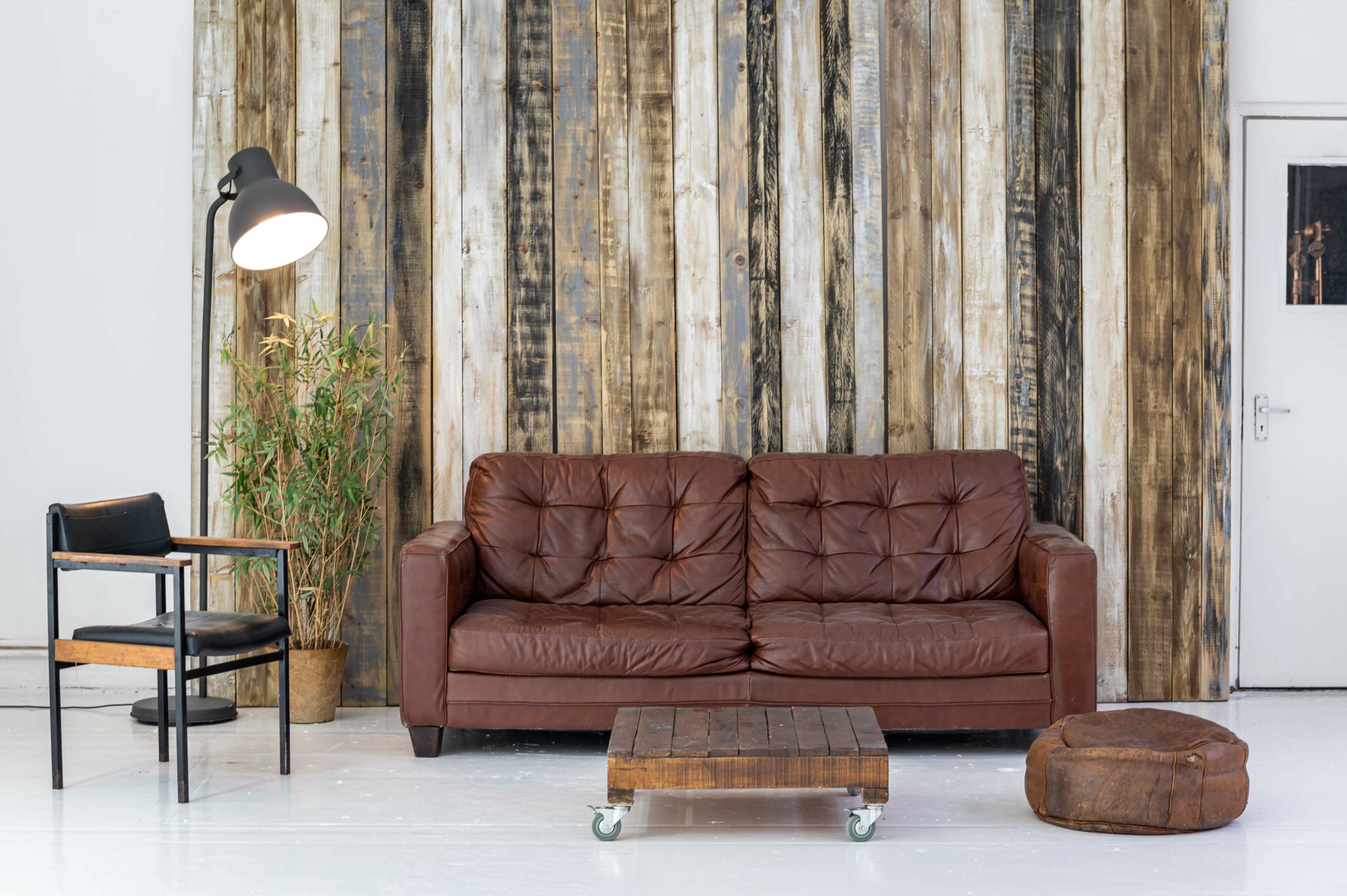 A brown leather sofa, a wooden coffee table on wheels, a black chair, and a potted plant against a backdrop of vertically arranged wooden planks.