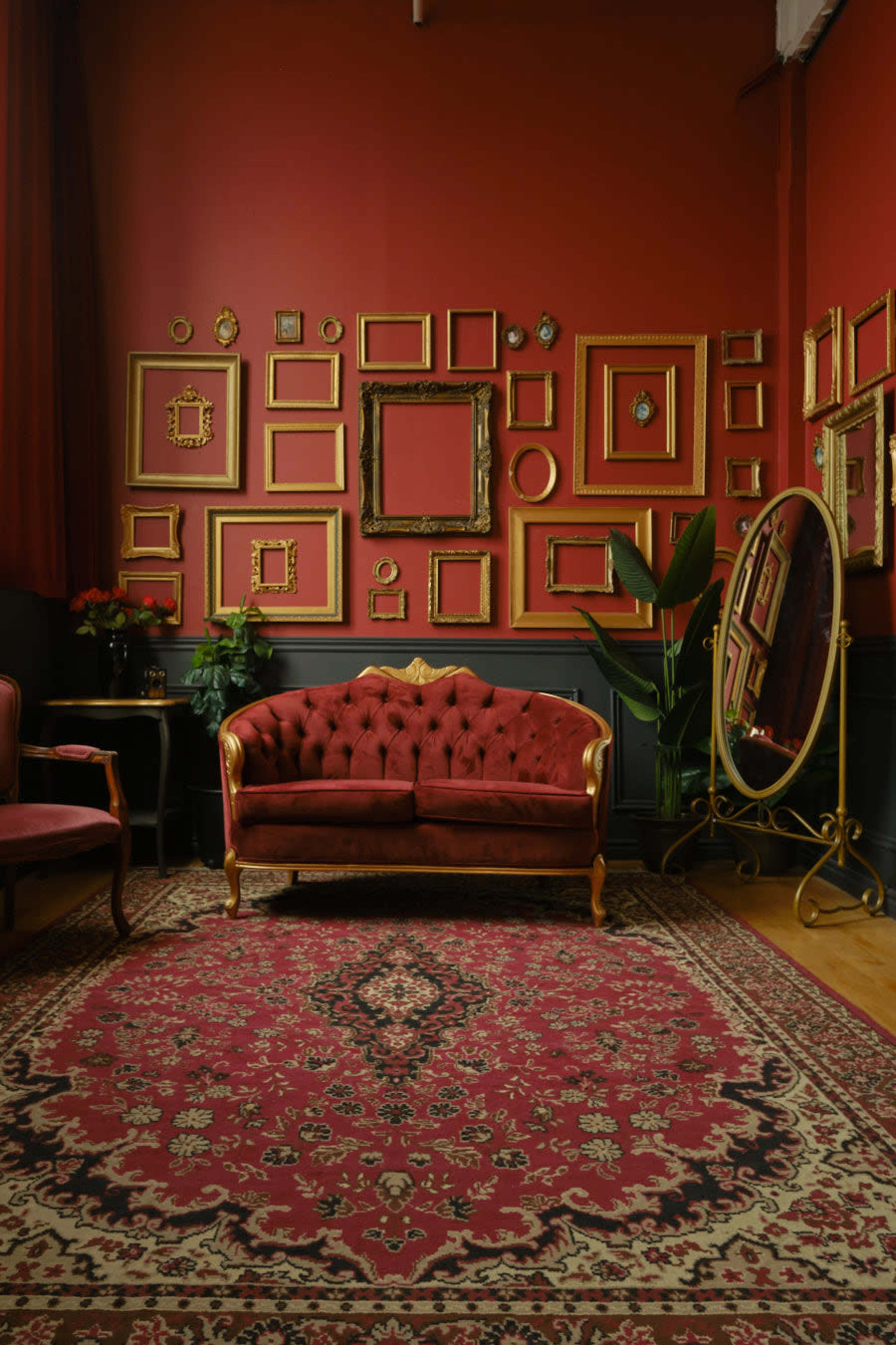A cozy living space featuring a red tufted sofa, an ornate mirror, and a collection of empty gold-framed paintings on a red wall.