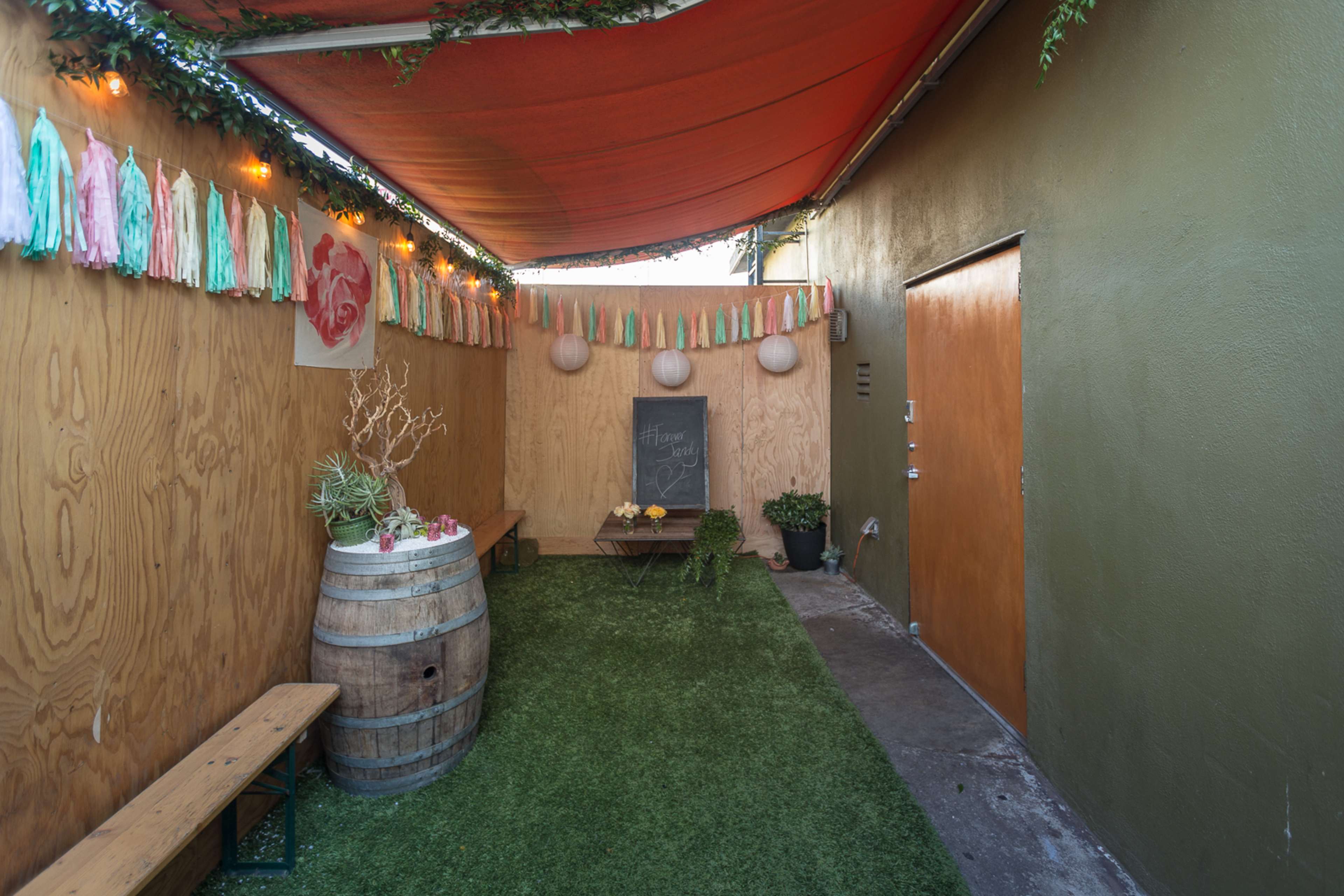 The image shows a narrow outdoor space with artificial grass, wooden walls, a barrel, a small table with flowers, and decorative hanging elements.