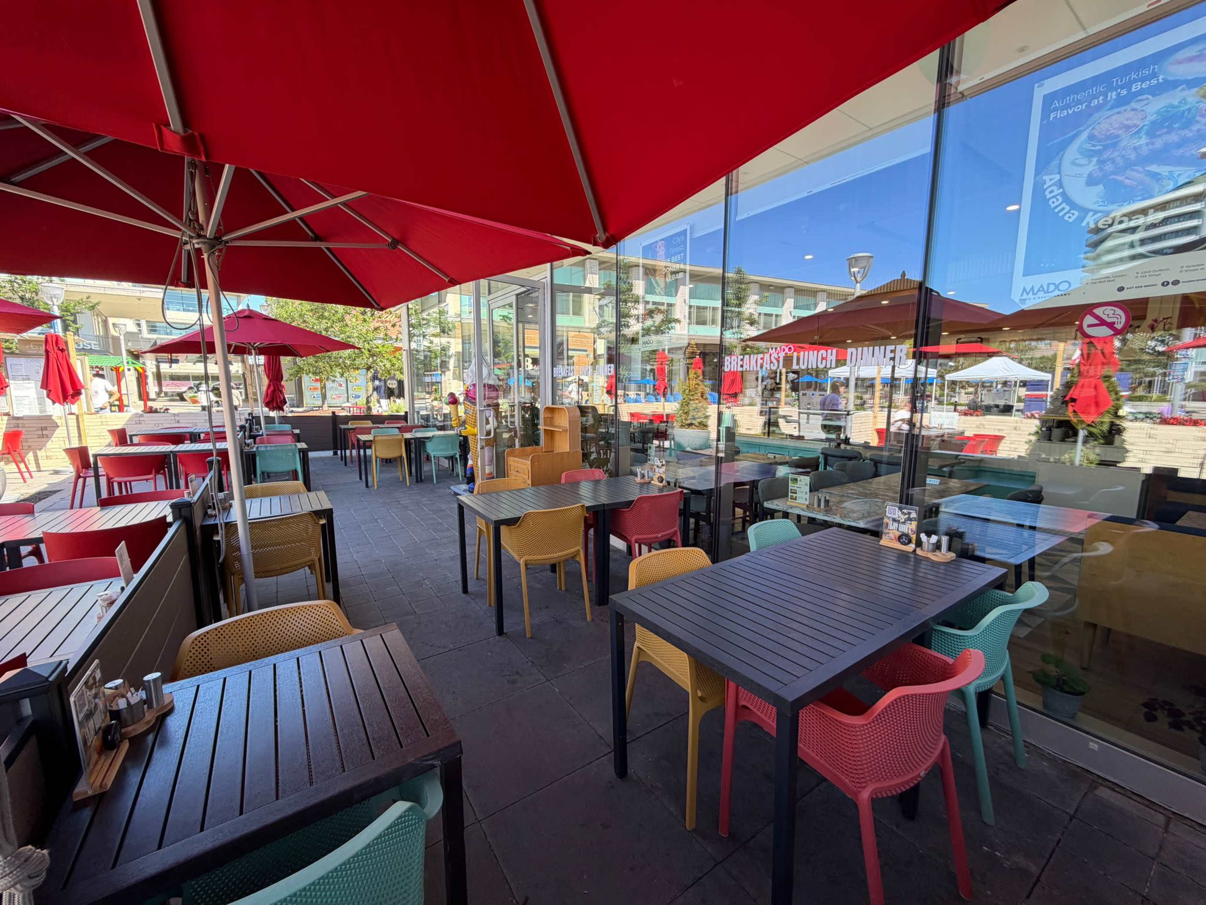 The scene shows an outdoor patio of a restaurant with colorful tables and chairs, shaded by large red umbrellas.
