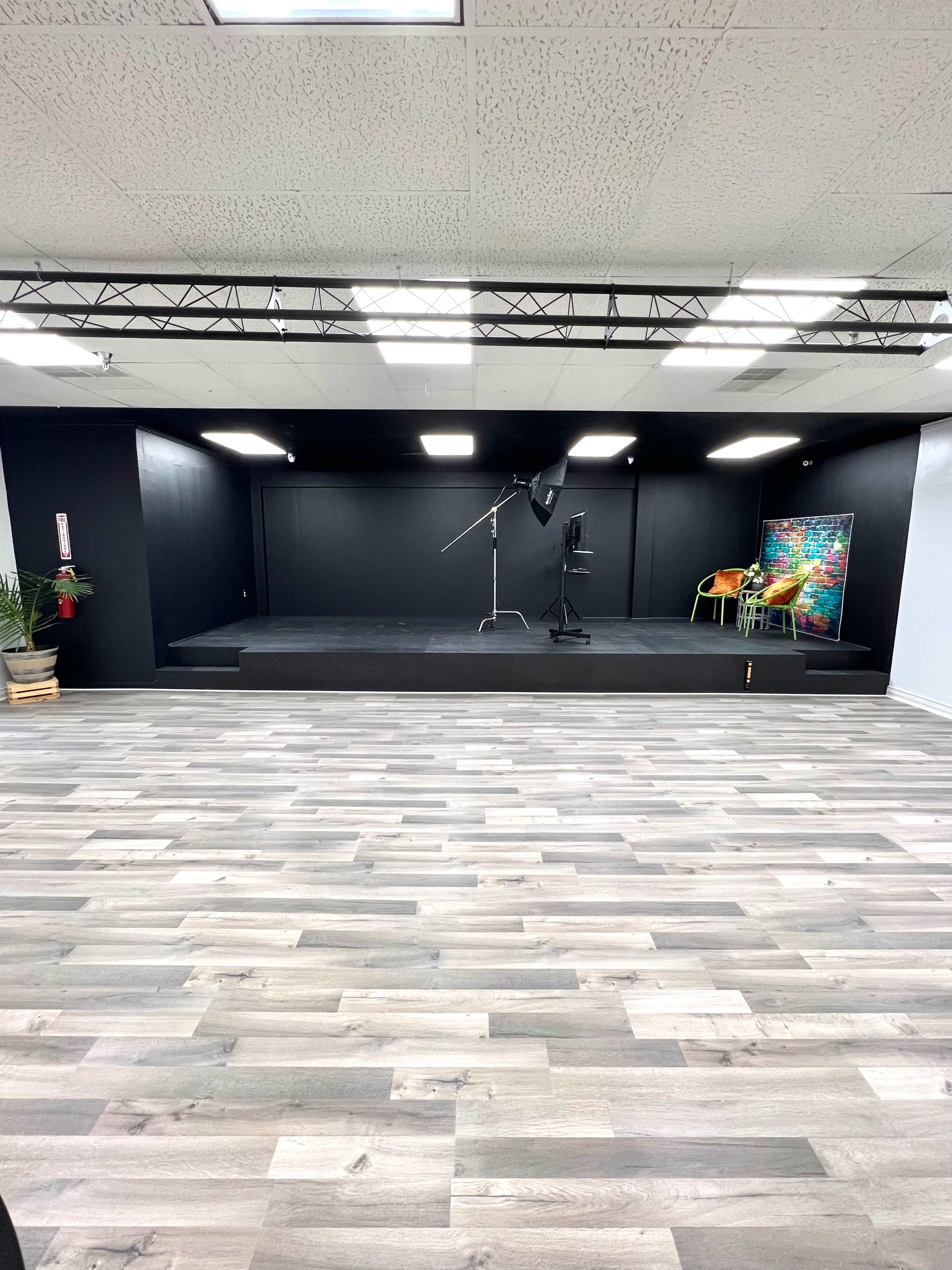 A stage with a black backdrop and lighting equipment in a room with gray wooden flooring and brightly colored artwork displayed to the side.