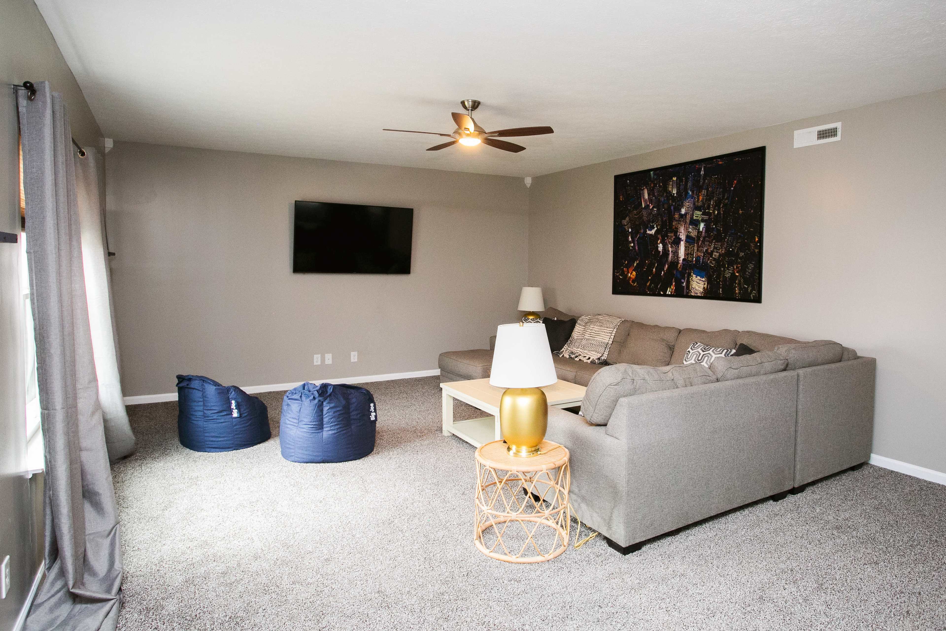 The living room features a gray sofa, a wall-mounted TV, a coffee table, two blue bean bags, and a lamp on a gold side table, all set against neutral walls and a carpeted floor.