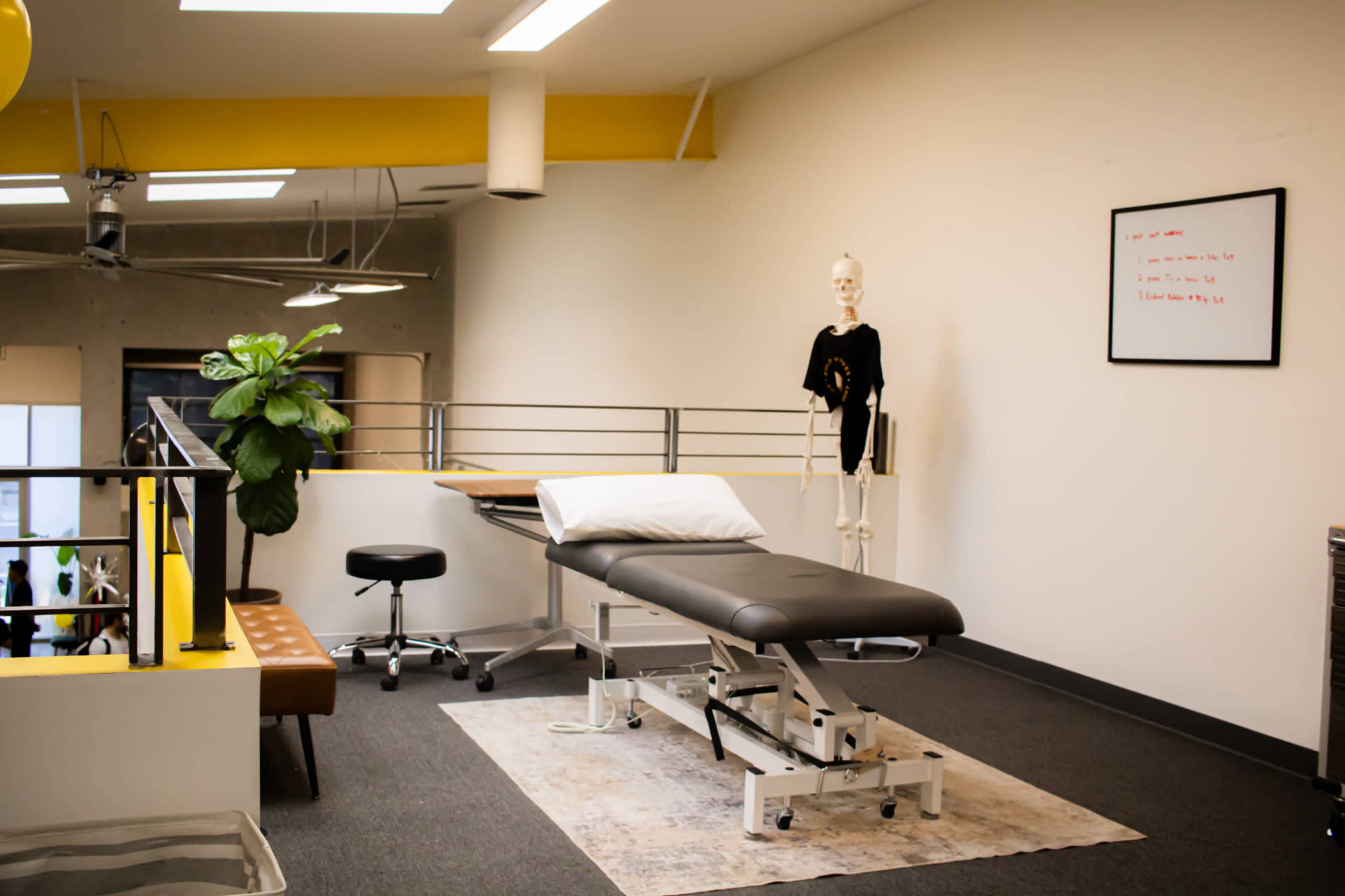 The image shows a treatment room featuring a massage table, medical skeleton model, office chair, and a plant, along with a whiteboard mounted on the wall.