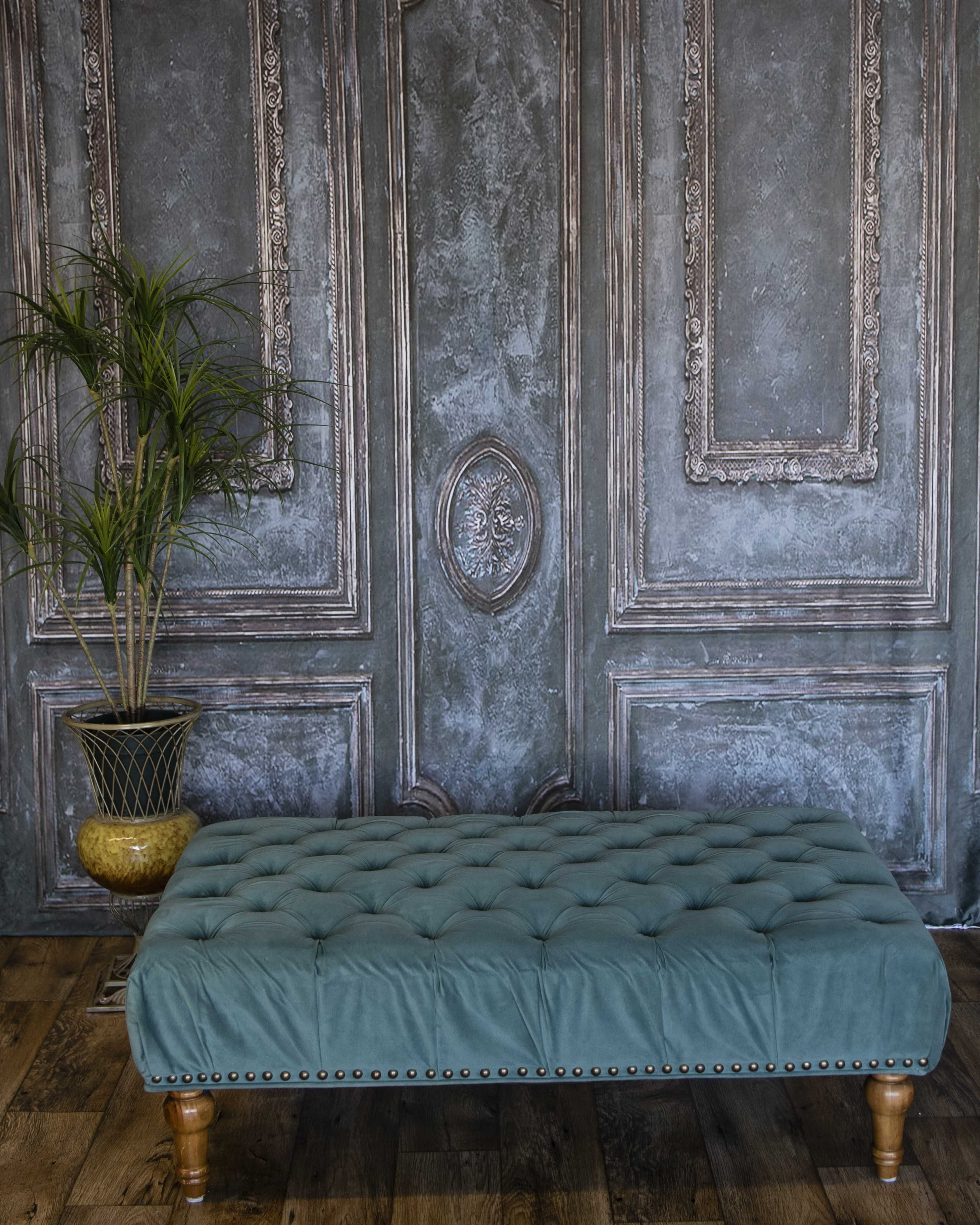 The image features a tufted teal upholstered bench placed against a textured gray wall with ornate paneling and a decorative plant in a pot beside it.