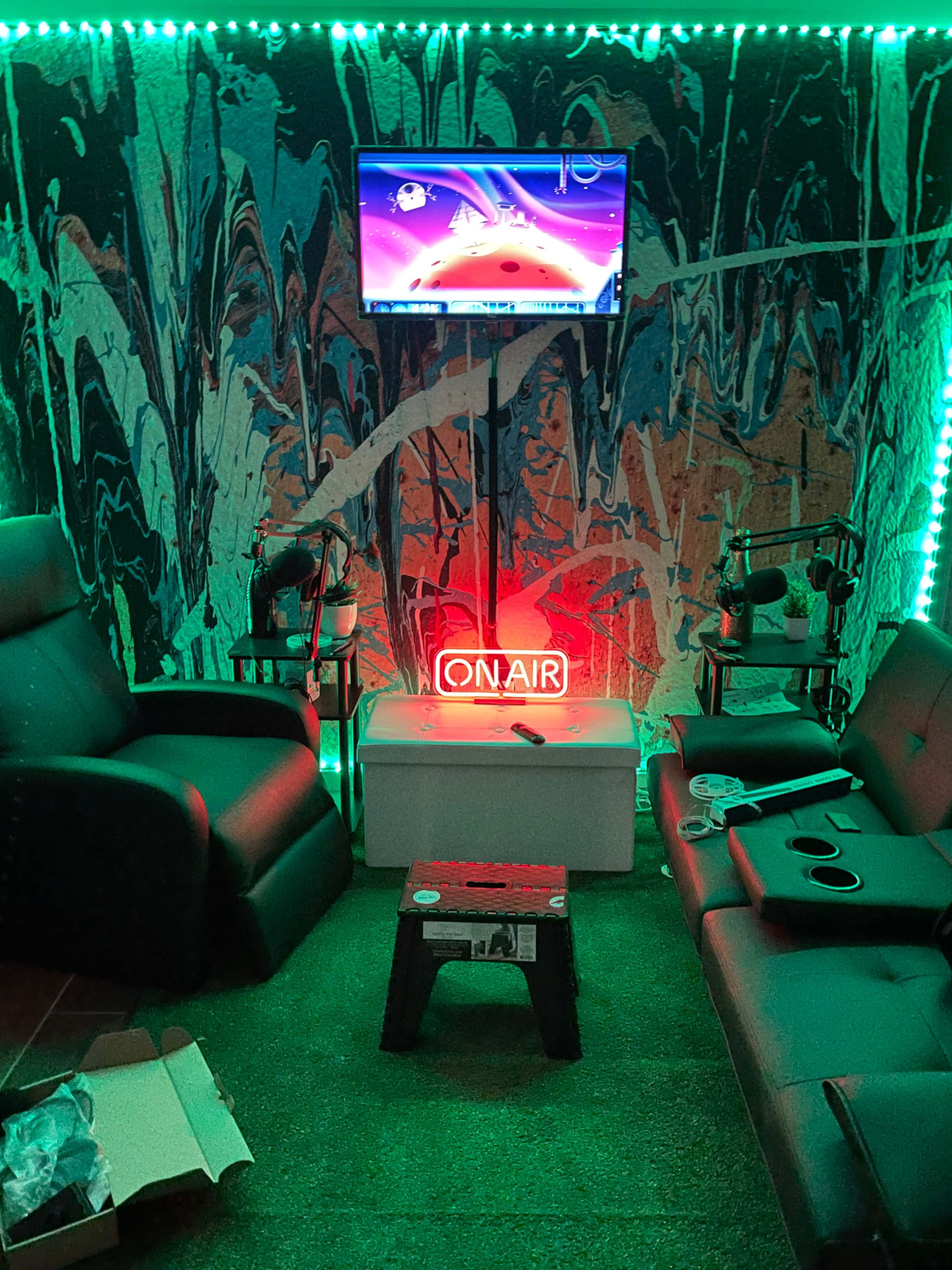 A room with two black couches facing a central table, a television mounted on the wall, and a neon "ON AIR" sign illuminated in red.