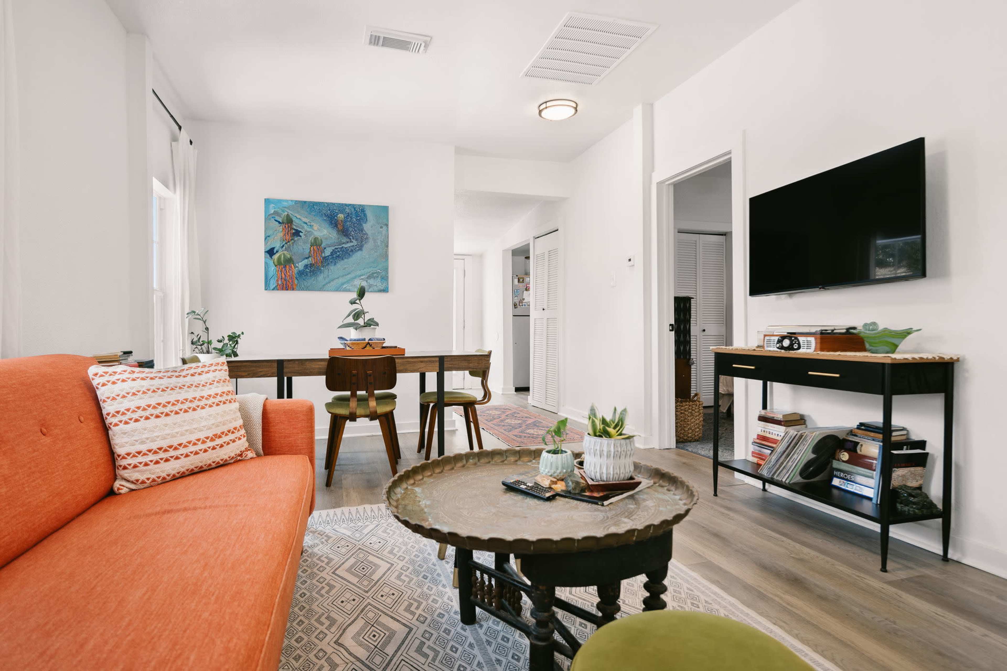 The image shows a living room featuring an orange couch, a wooden table with chairs, a TV stand with a television, and various decorative elements in a bright, modern space.