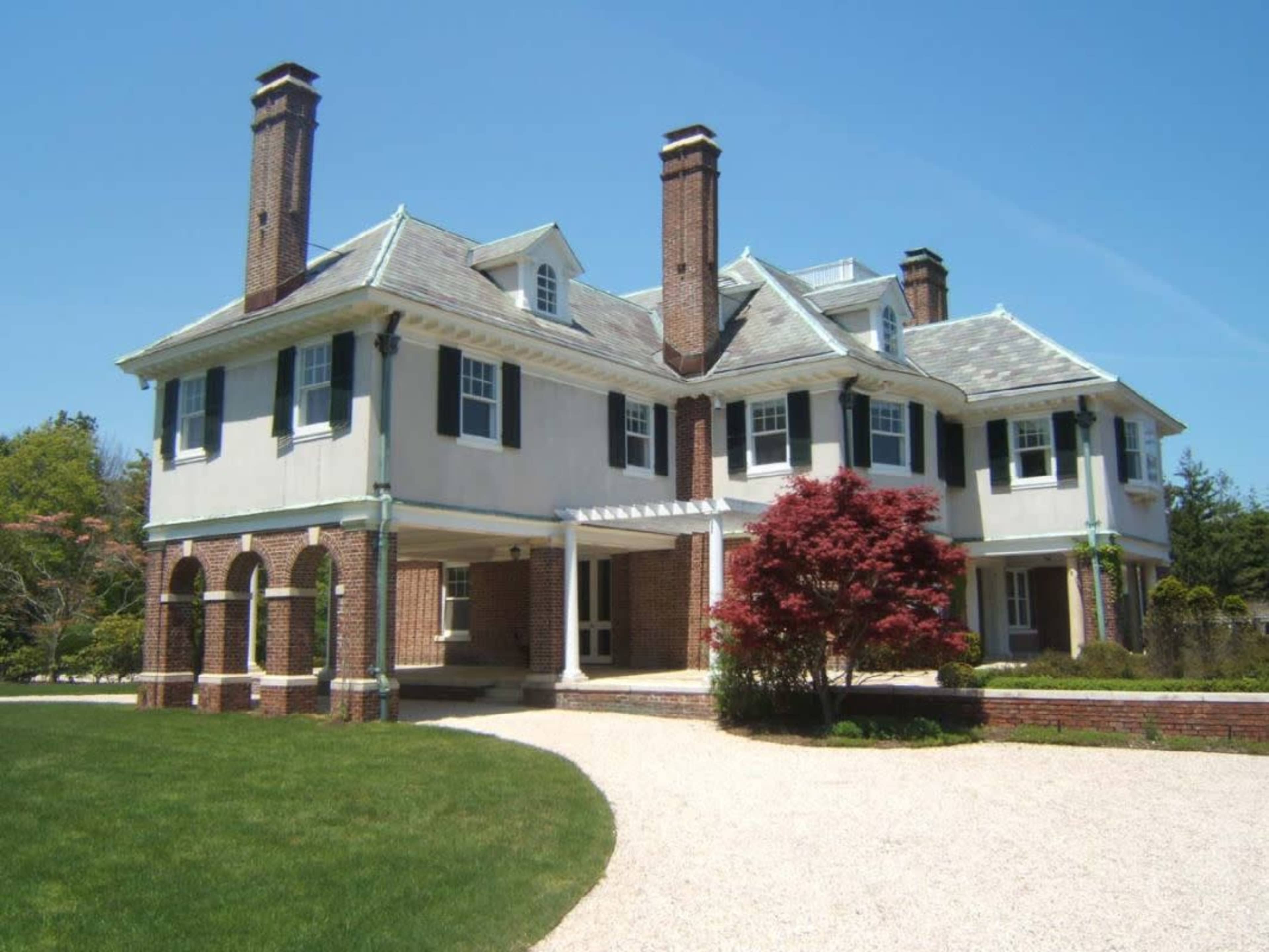 The image shows a large, two-story mansion with a circular driveway, visible brick chimneys, and a landscaped front yard.