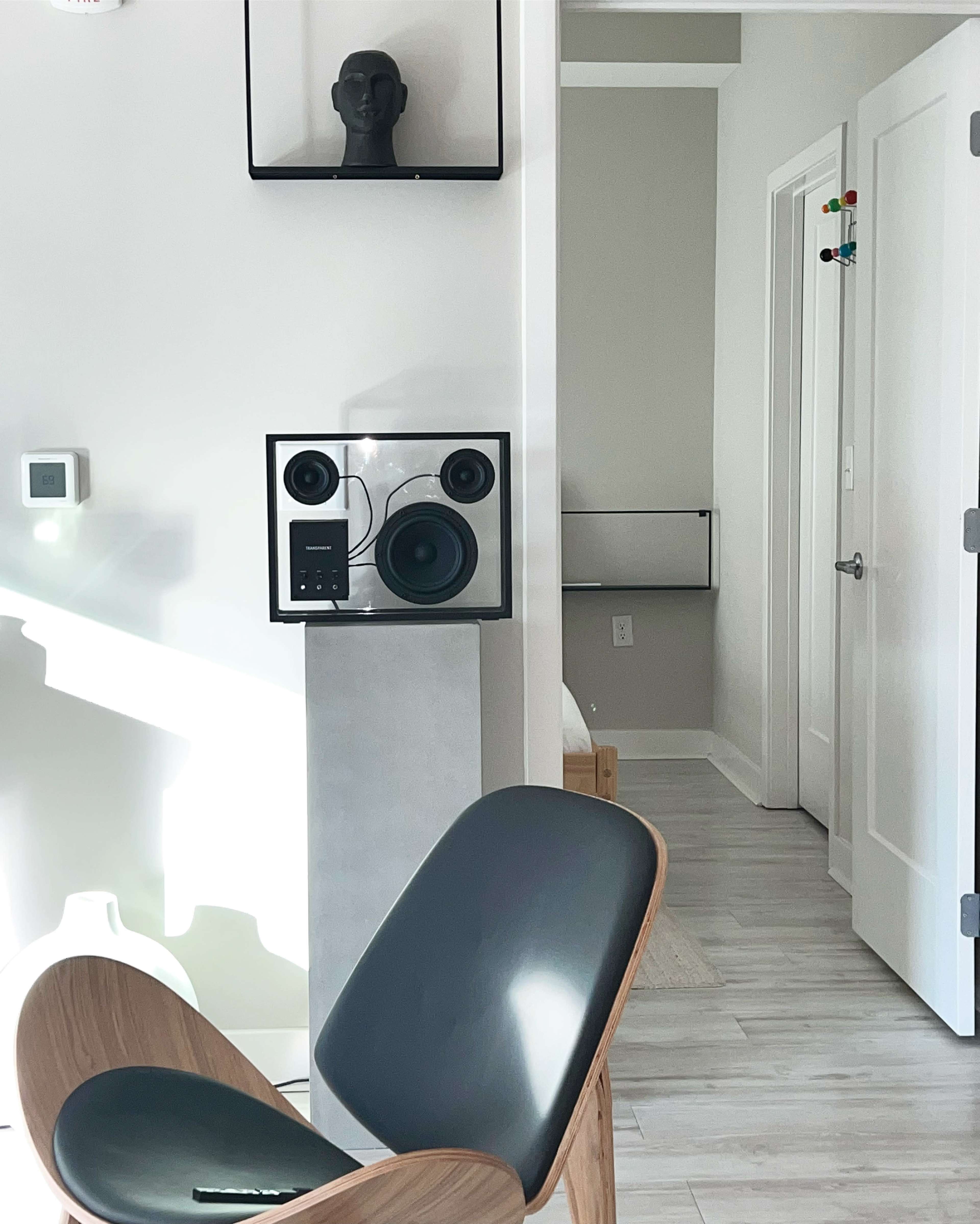 A modern interior with a wooden chair, a speaker on a stand, and a minimalist décor that includes a framed sculpture on the wall.