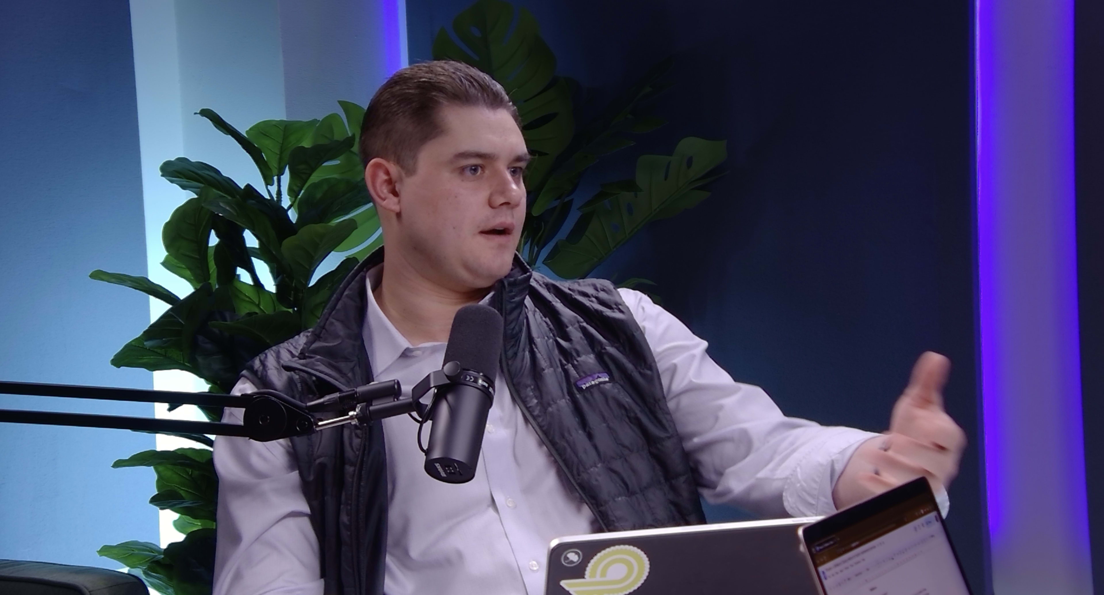 A man wearing a white shirt and black vest sits at a table with a microphone and a laptop, gesturing while speaking.