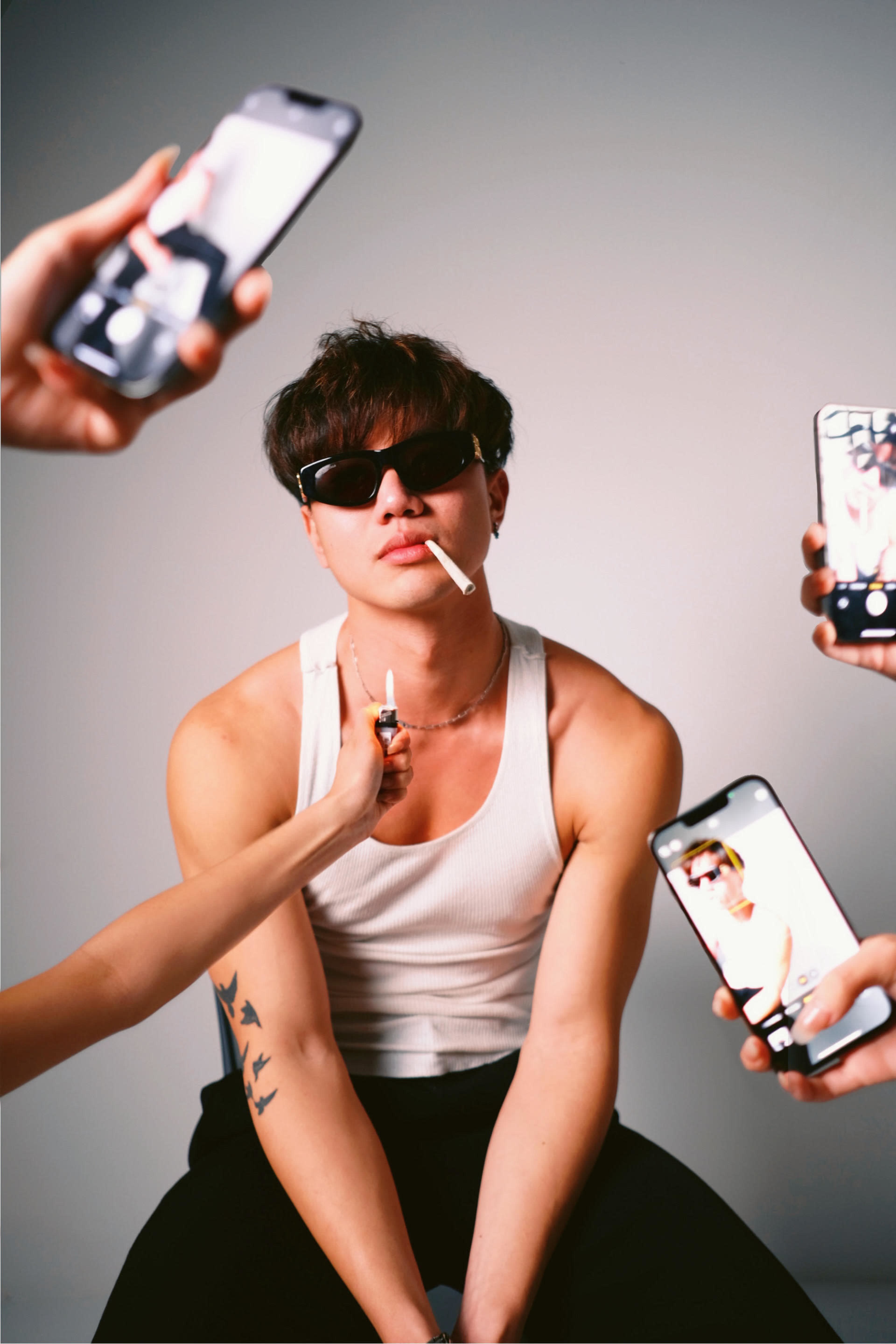 A young man wearing sunglasses and a tank top poses with a cigarette, while several hands hold smartphones recording him.