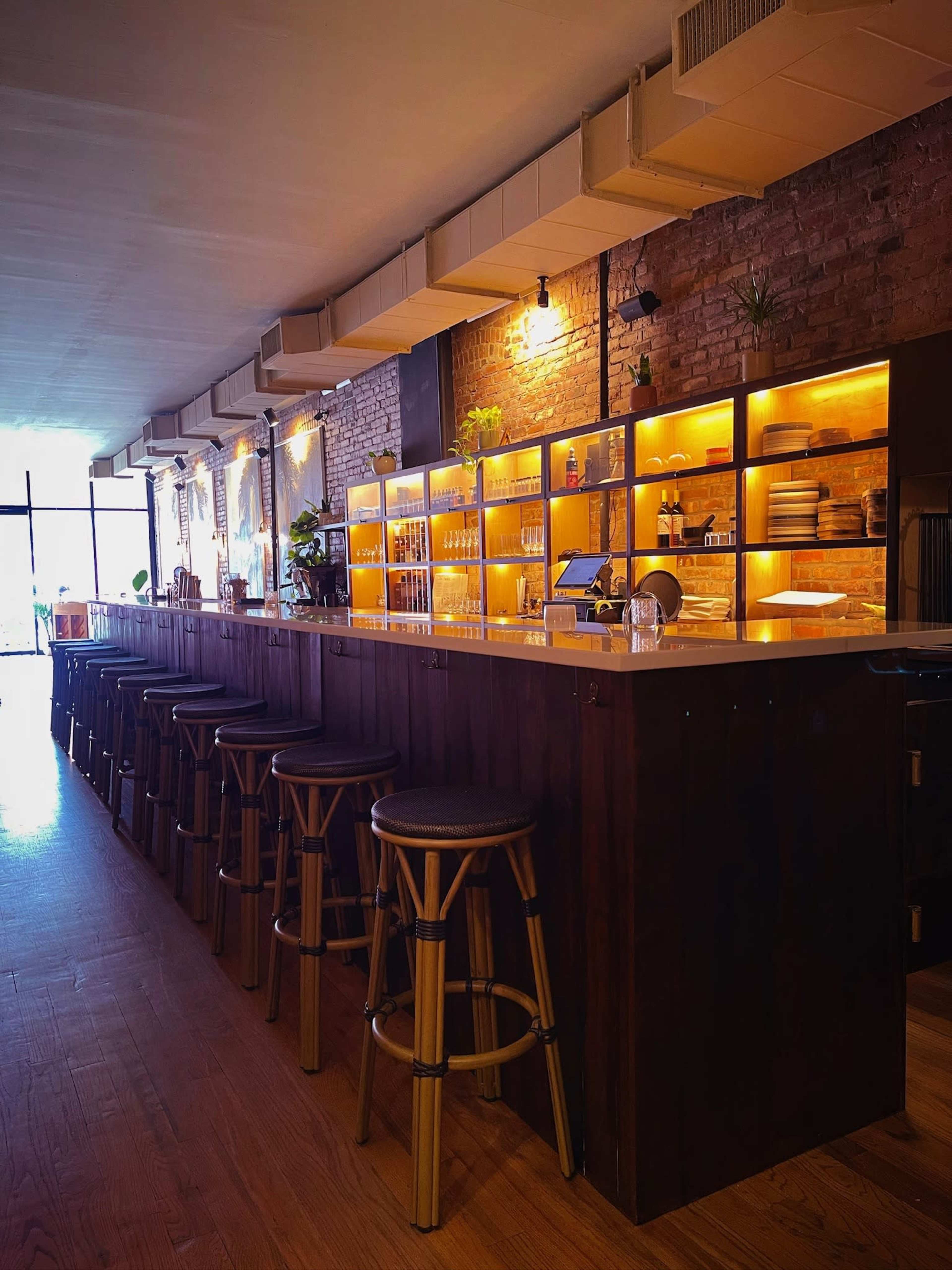 The image shows a modern bar with wooden stools, illuminated shelves, and a warm atmosphere created by lighting along a brick wall.