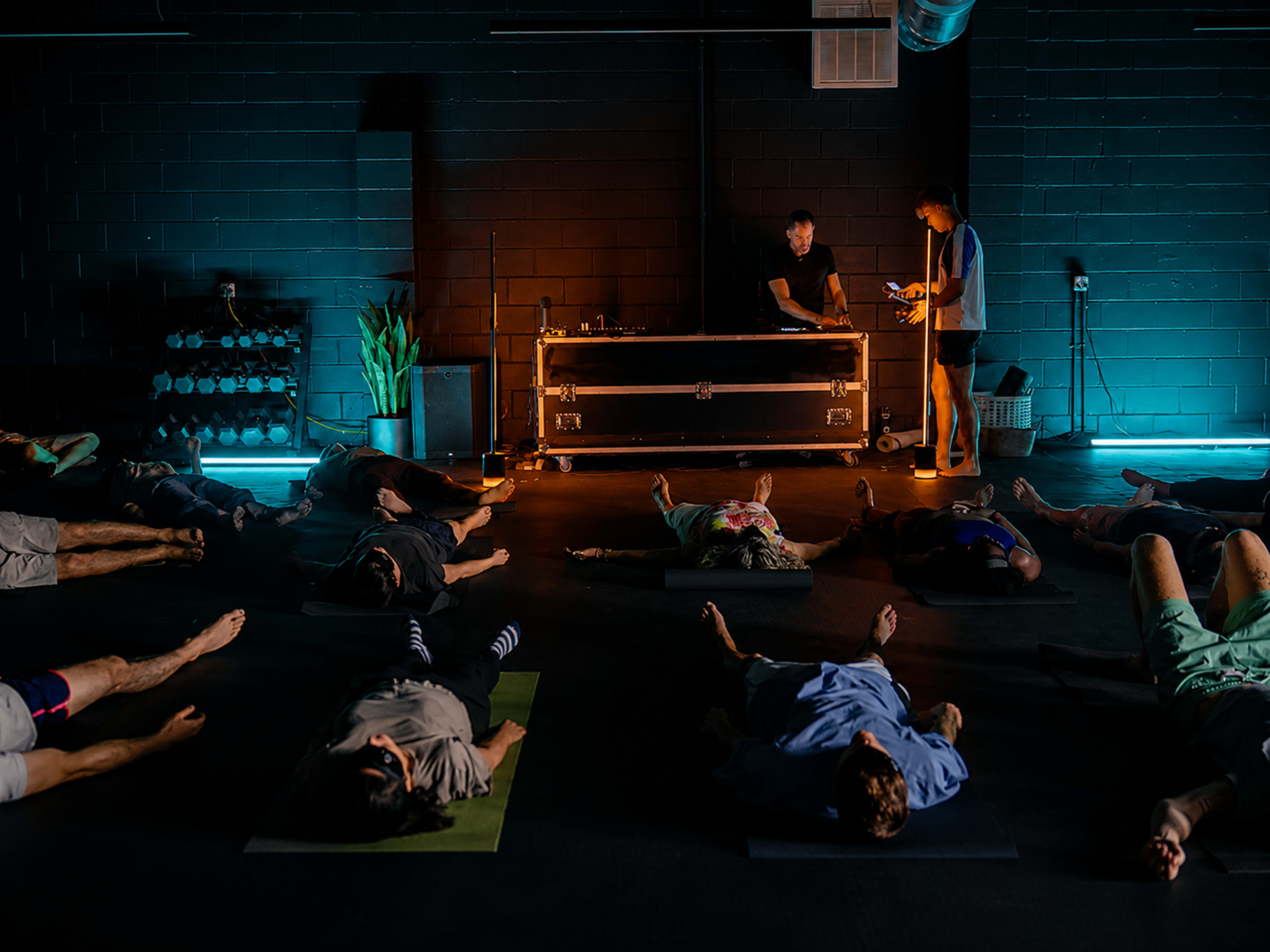 A group of people lies on yoga mats in a dimly lit room while two instructors stand by a table with equipment at the front.