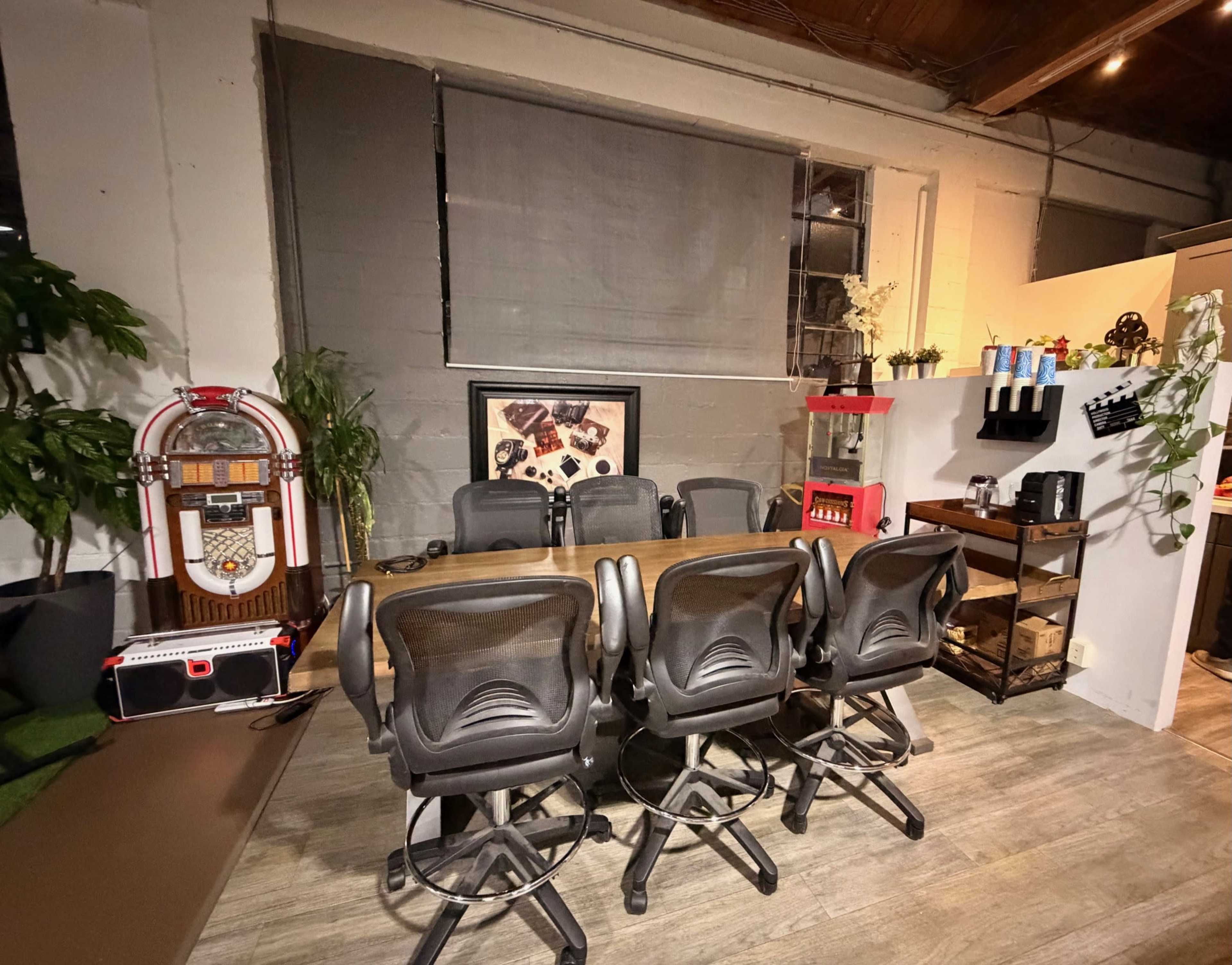 The image shows a modern office space with a large table surrounded by ergonomic chairs, a jukebox in the corner, and shelves displaying office supplies.