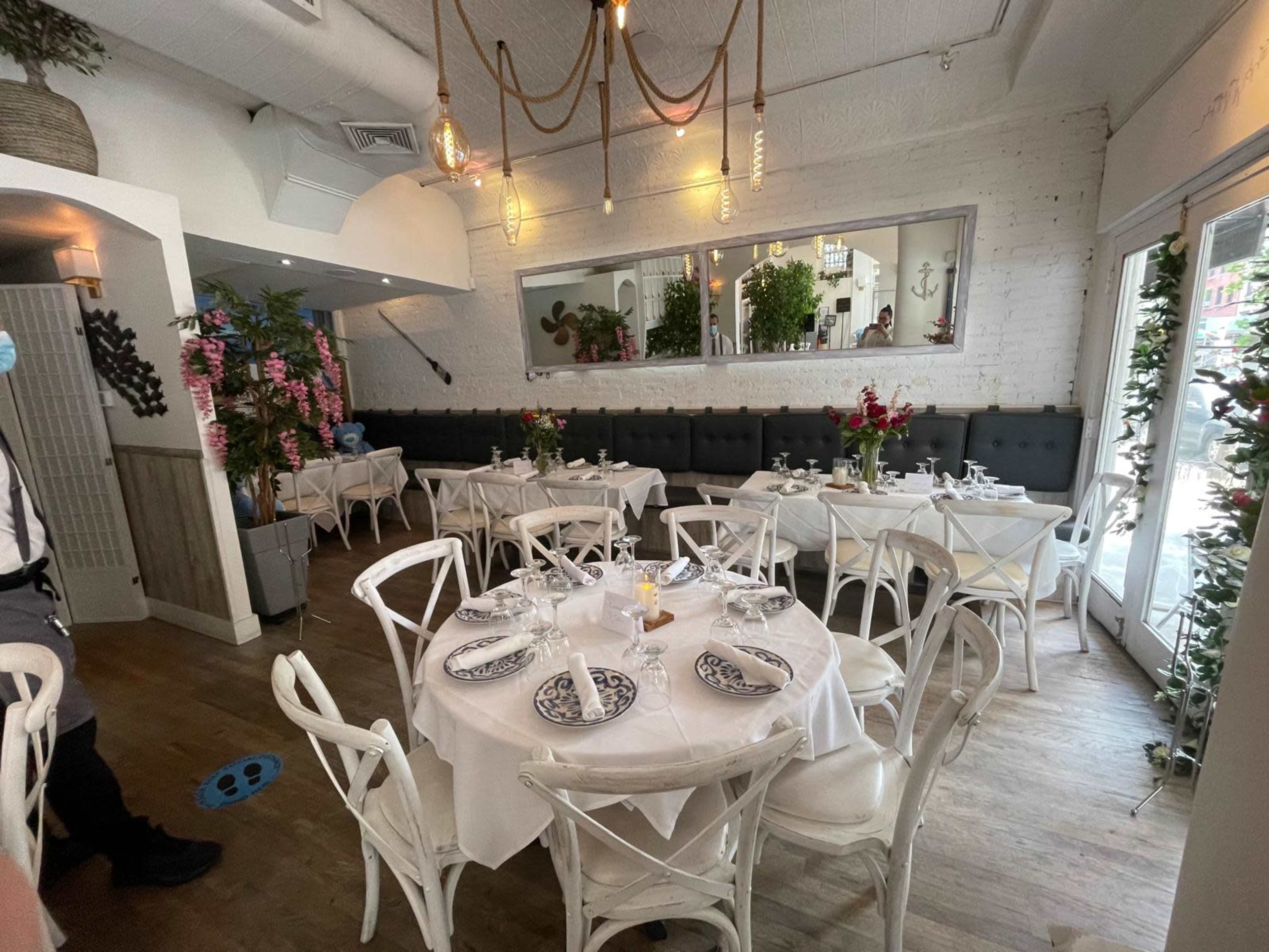 The image shows a restaurant interior featuring elegantly set tables with white tablecloths and decorative plates, surrounded by floral arrangements and a large mirror on the wall.