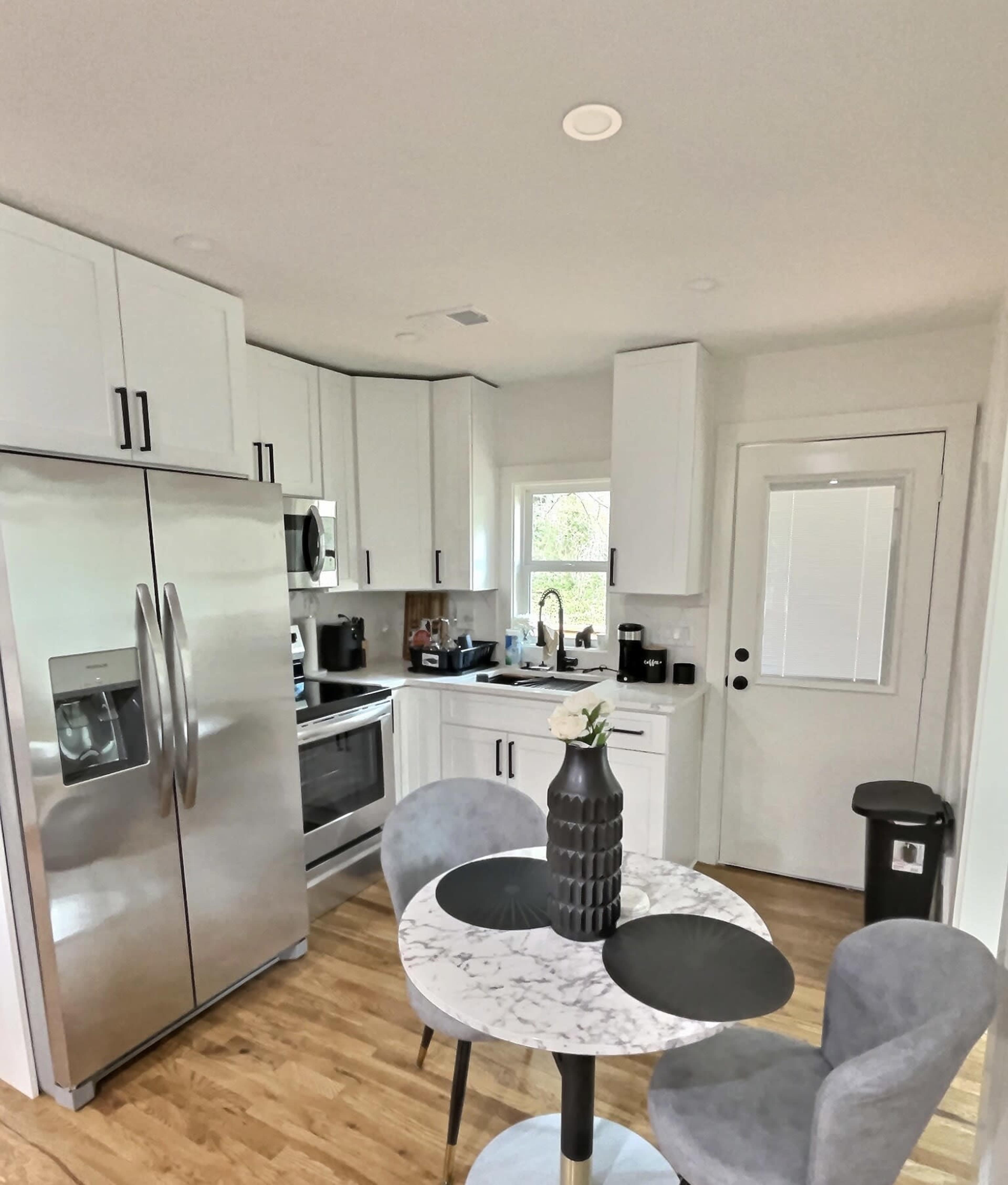 A modern kitchen featuring stainless steel appliances, a round marble dining table with two chairs, and a window providing natural light.
