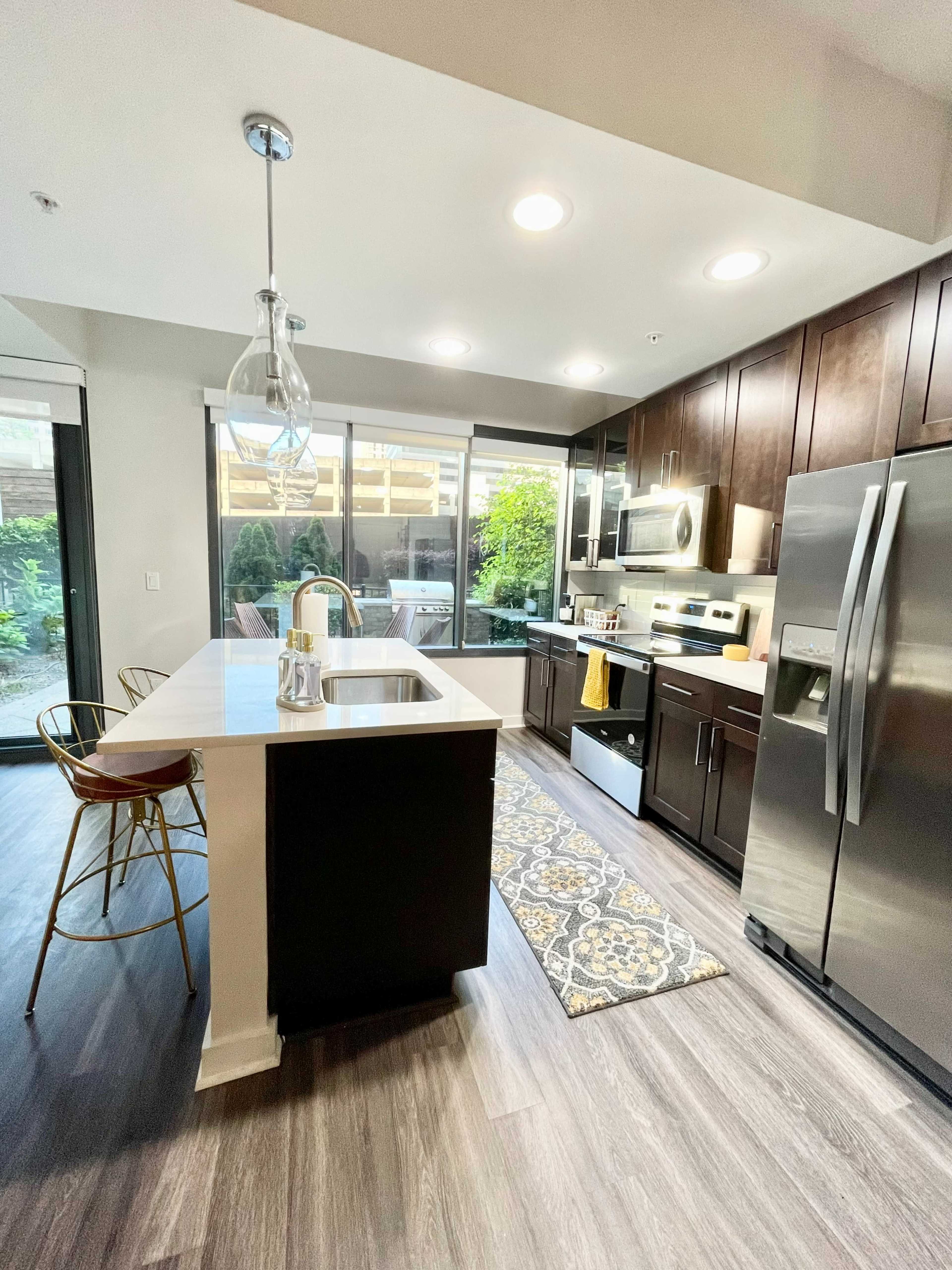 A modern kitchen features dark cabinetry, stainless steel appliances, a large island with seating, and a view of an outdoor patio through a glass door.