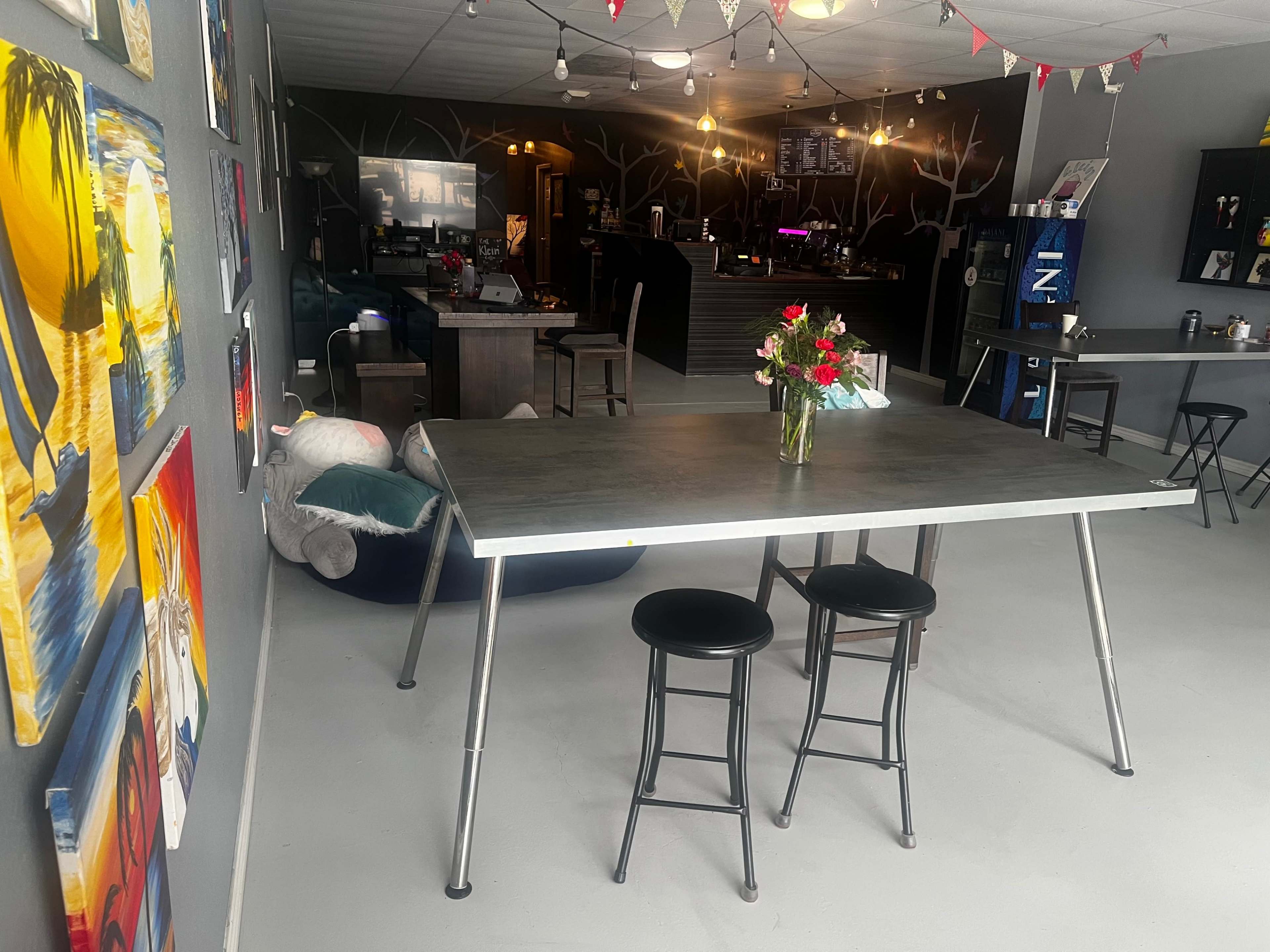 A modern dining table with two black stools stands in a room decorated with colorful paintings, leading to a bar area in the background.