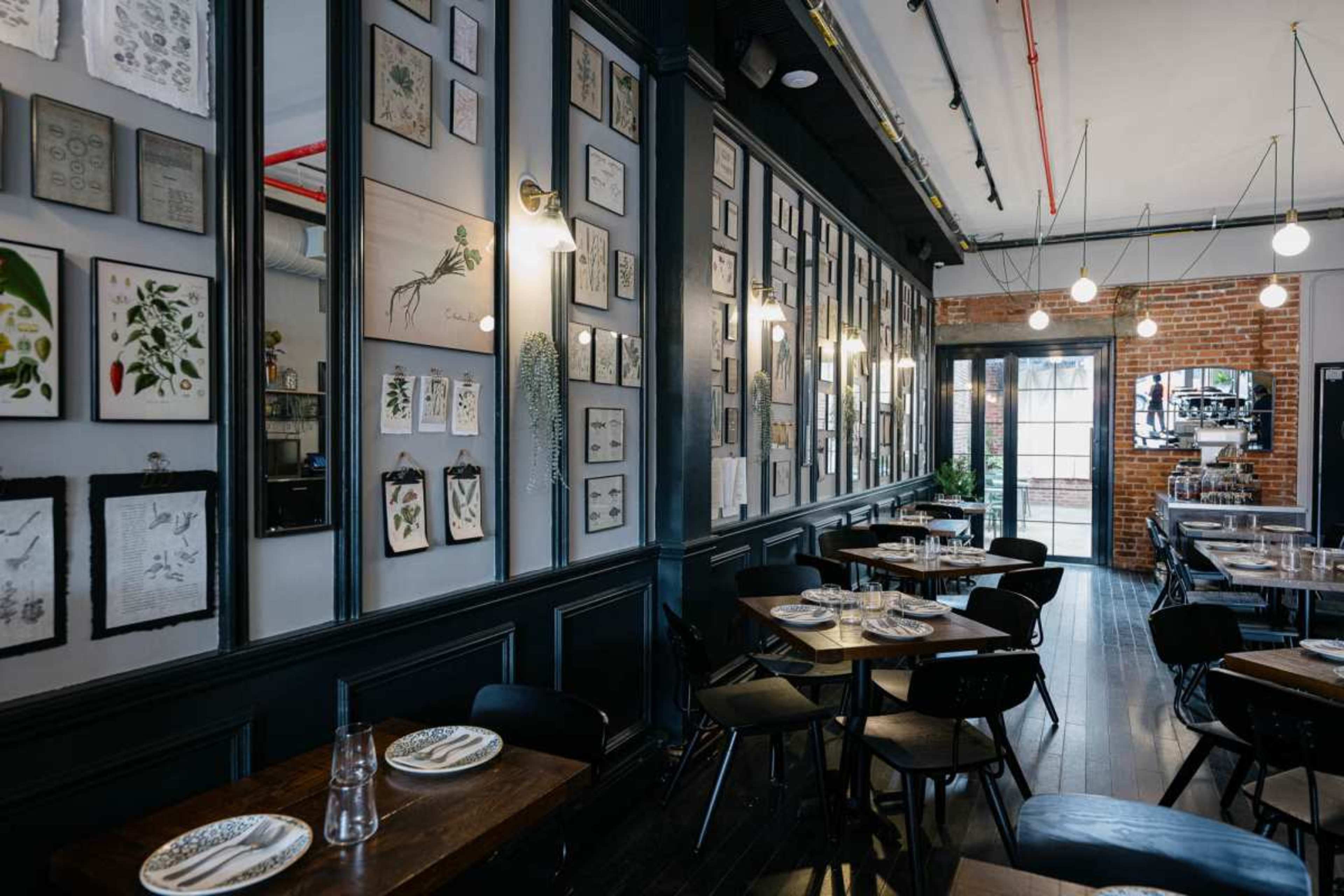 The image shows the interior of a restaurant with framed artwork on the walls, wooden tables set for dining, and modern lighting fixtures.