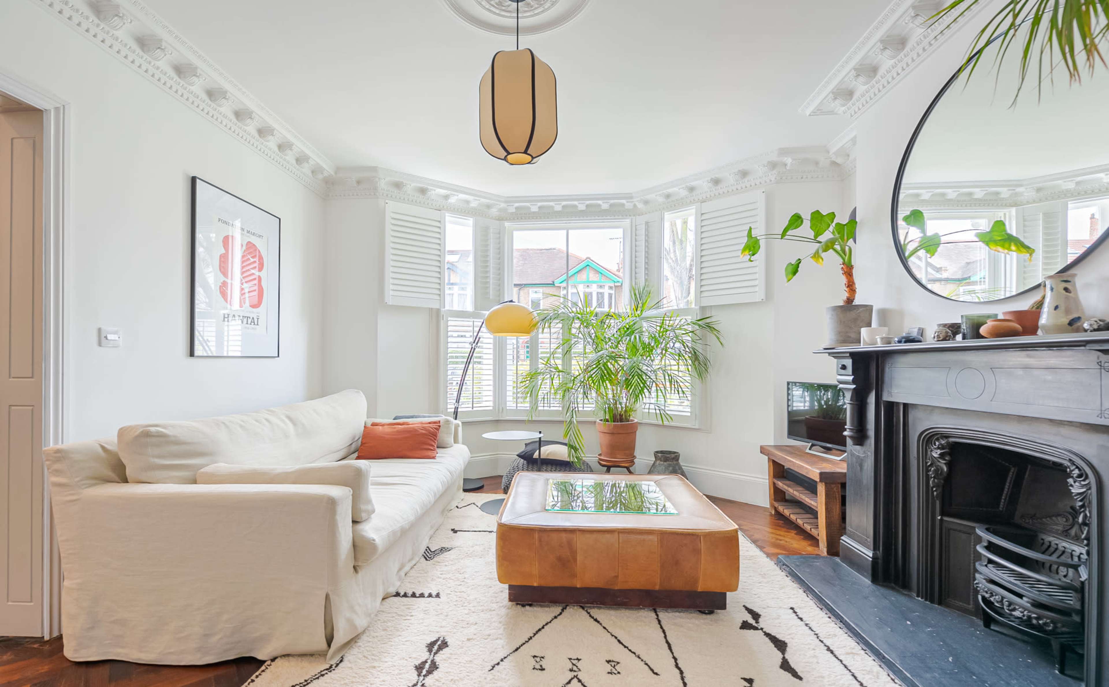 A living room features a white sofa, a large coffee table, and green plants, with bay windows and decorative molding.