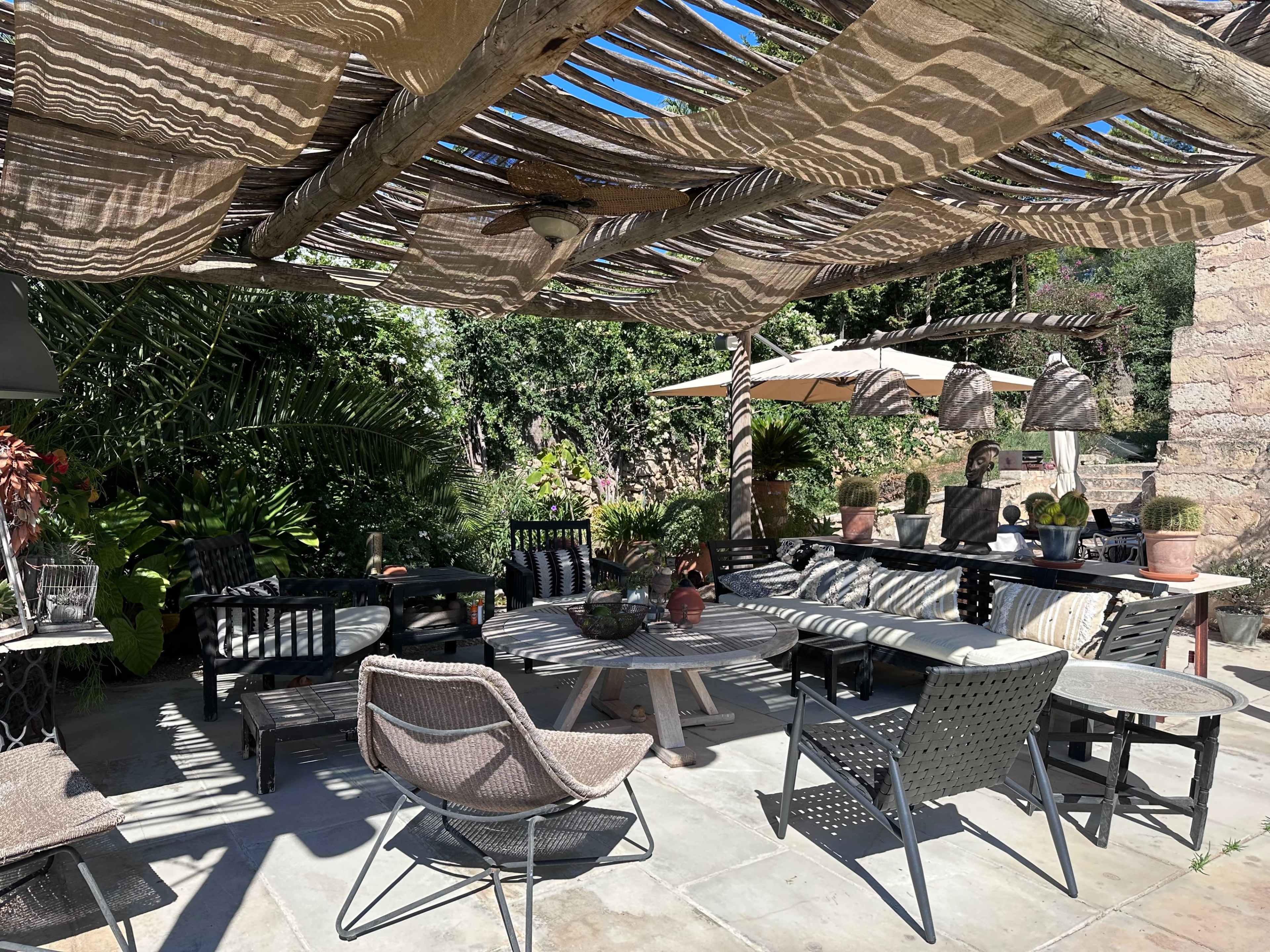 A shaded outdoor seating area features various chairs and a table, surrounded by green plants and decorative elements.