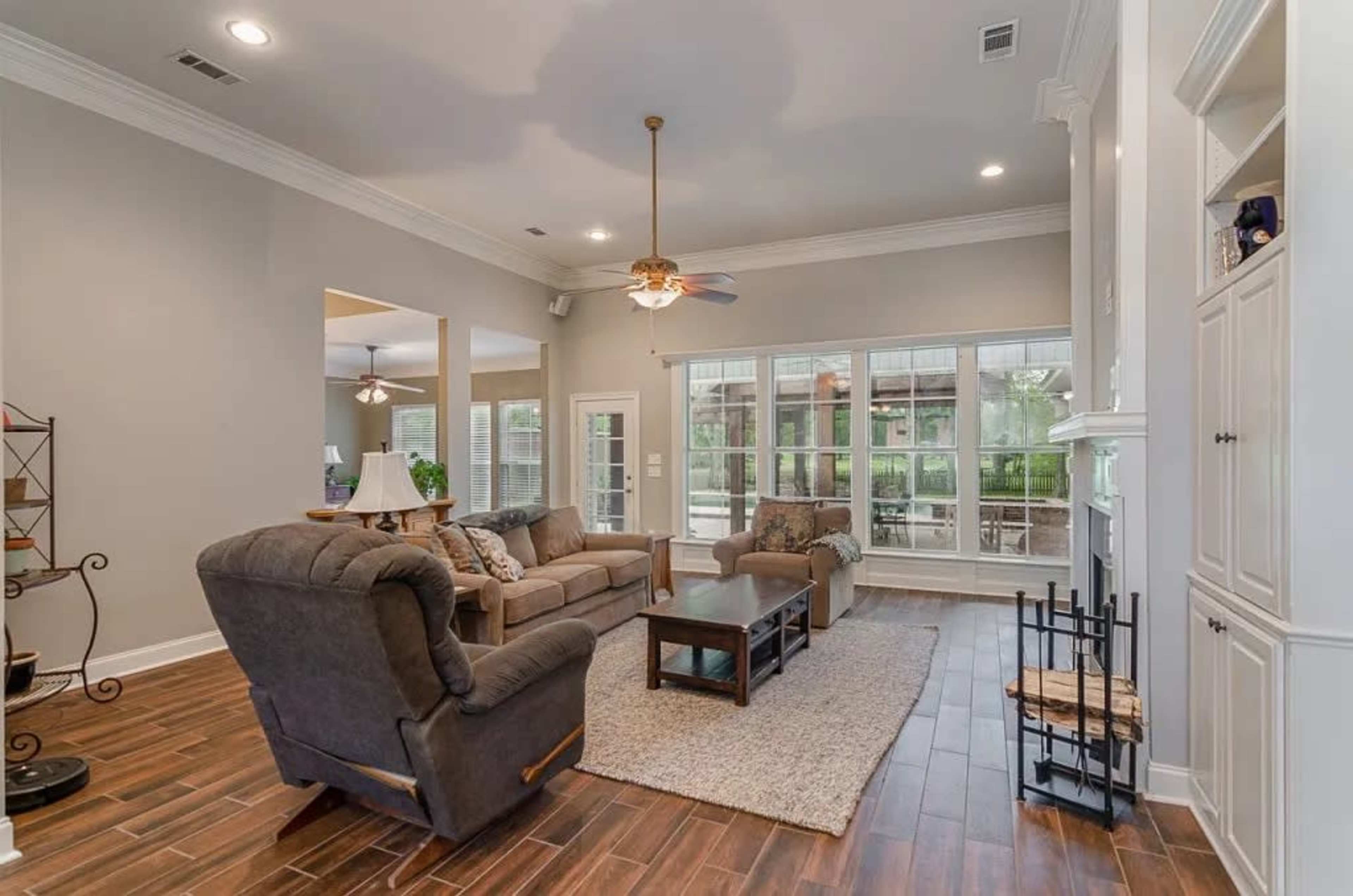 The image shows a spacious living room with a ceiling fan, large windows, and comfortable seating around a coffee table.