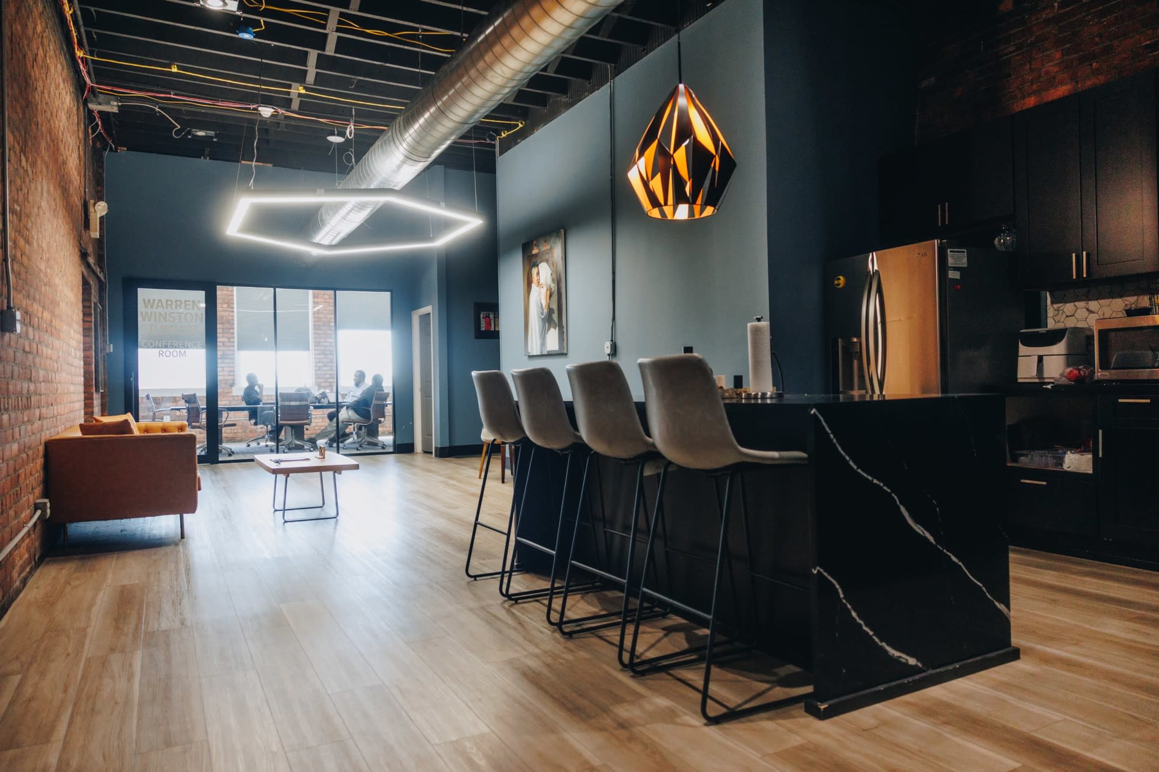 The image shows a modern kitchen area with a black granite island and bar stools, adjacent to a bright seating area and a workspace in the background.