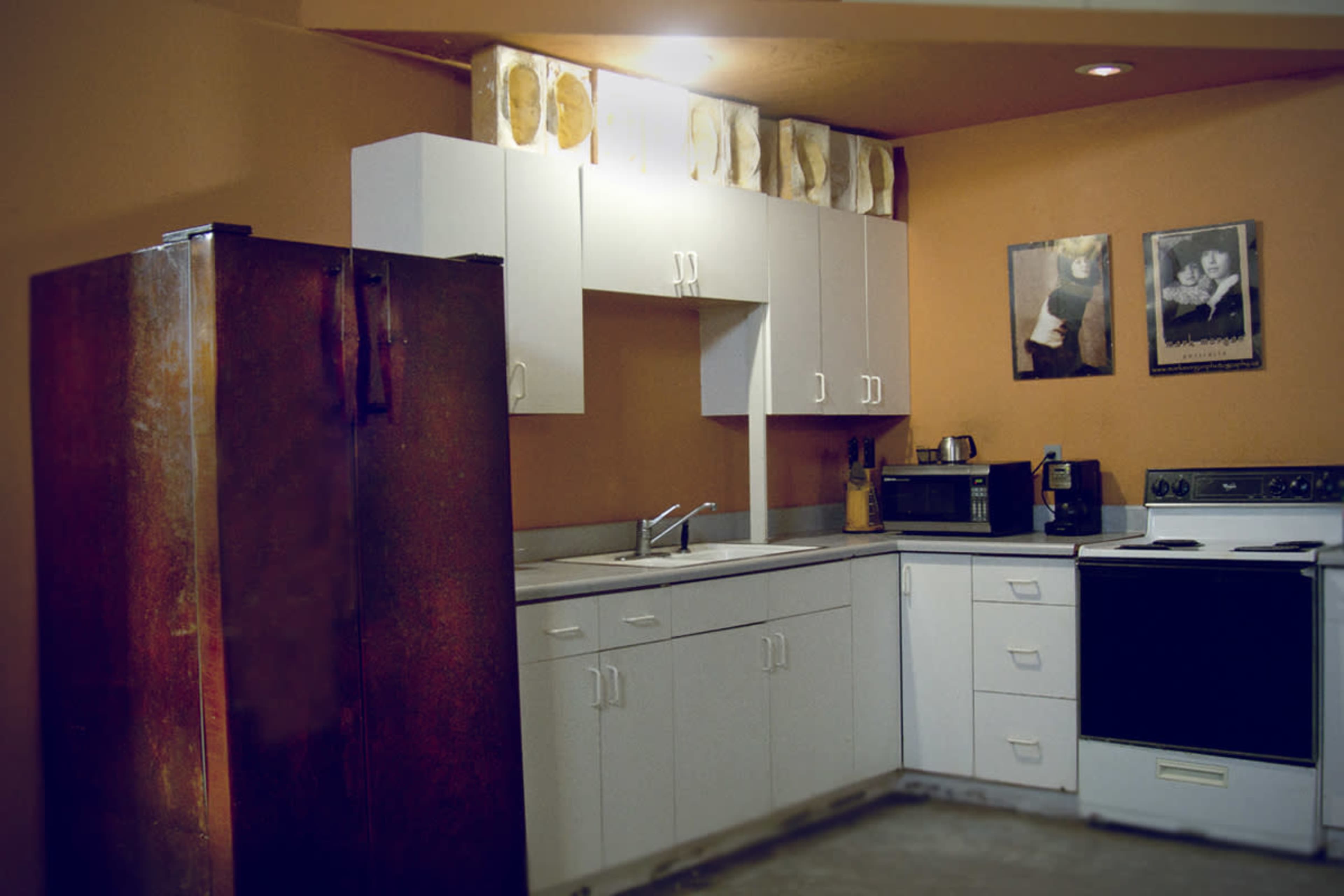 A kitchen with white cabinets and appliances, featuring a stainless steel refrigerator, a stove, and a sink, against an orange-painted wall.