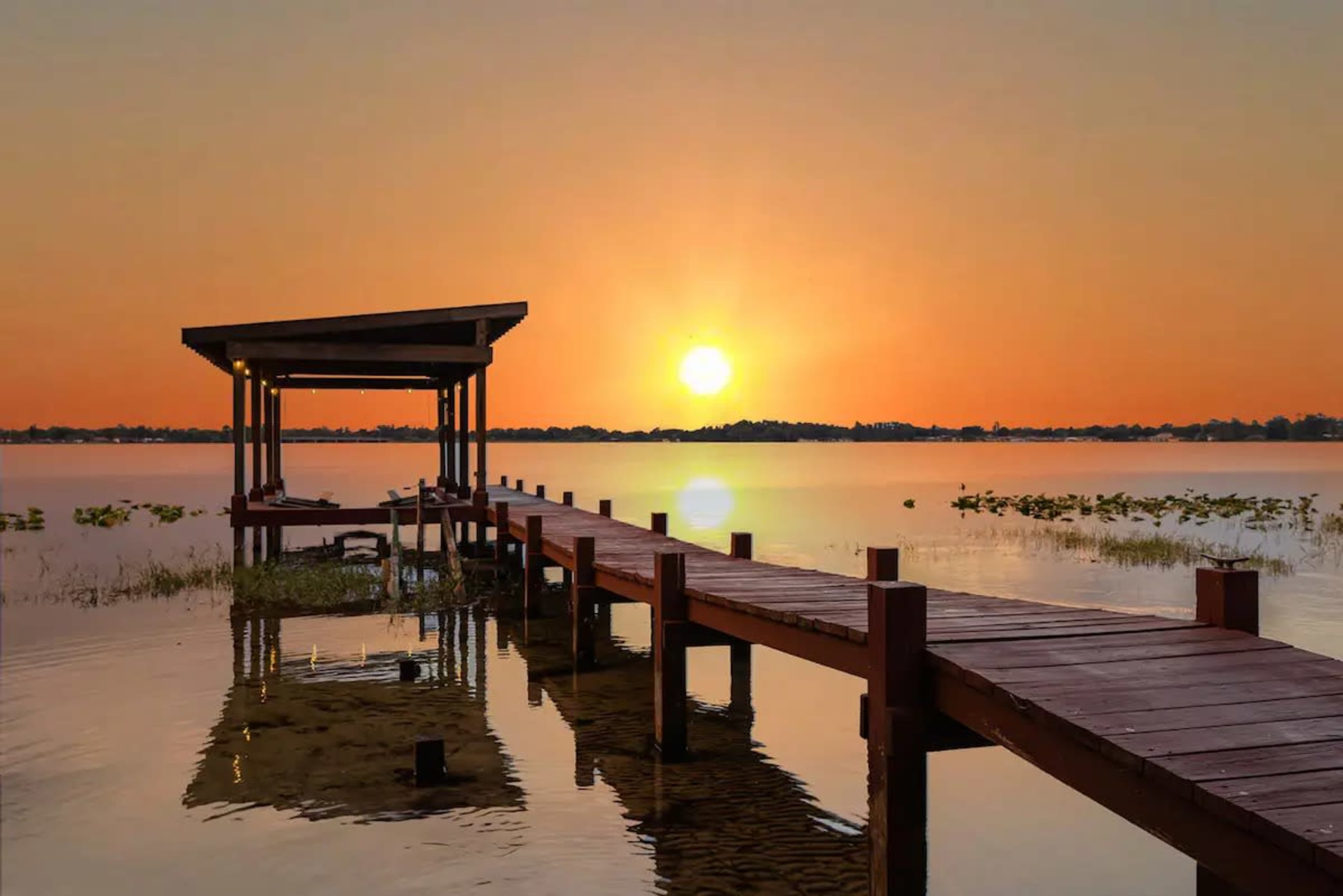 A wooden dock extends over a calm lake, leading to a covered structure as the sun sets on the horizon.