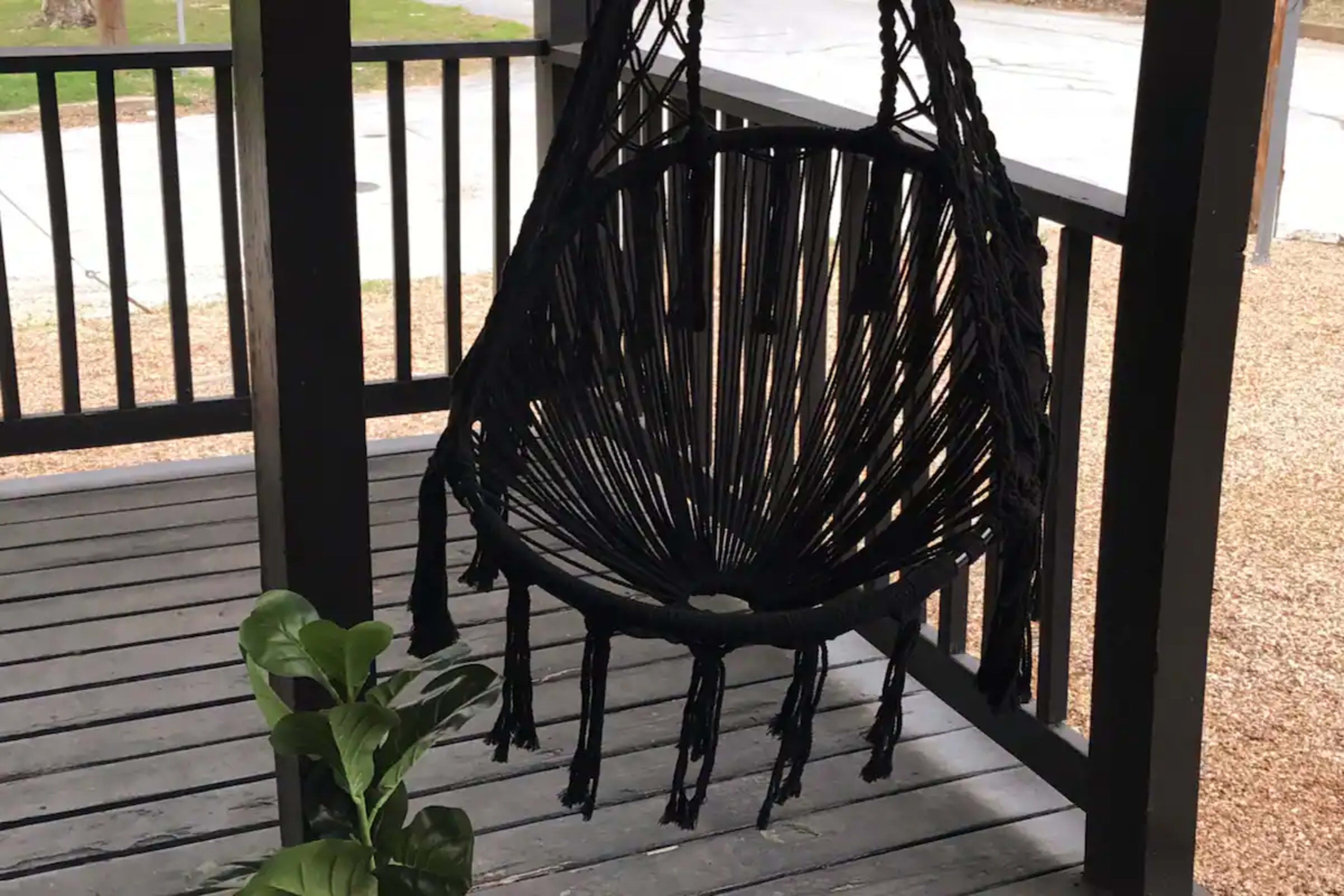 A black macramé hanging chair is suspended from the ceiling of a porch, with a potted plant beside it.