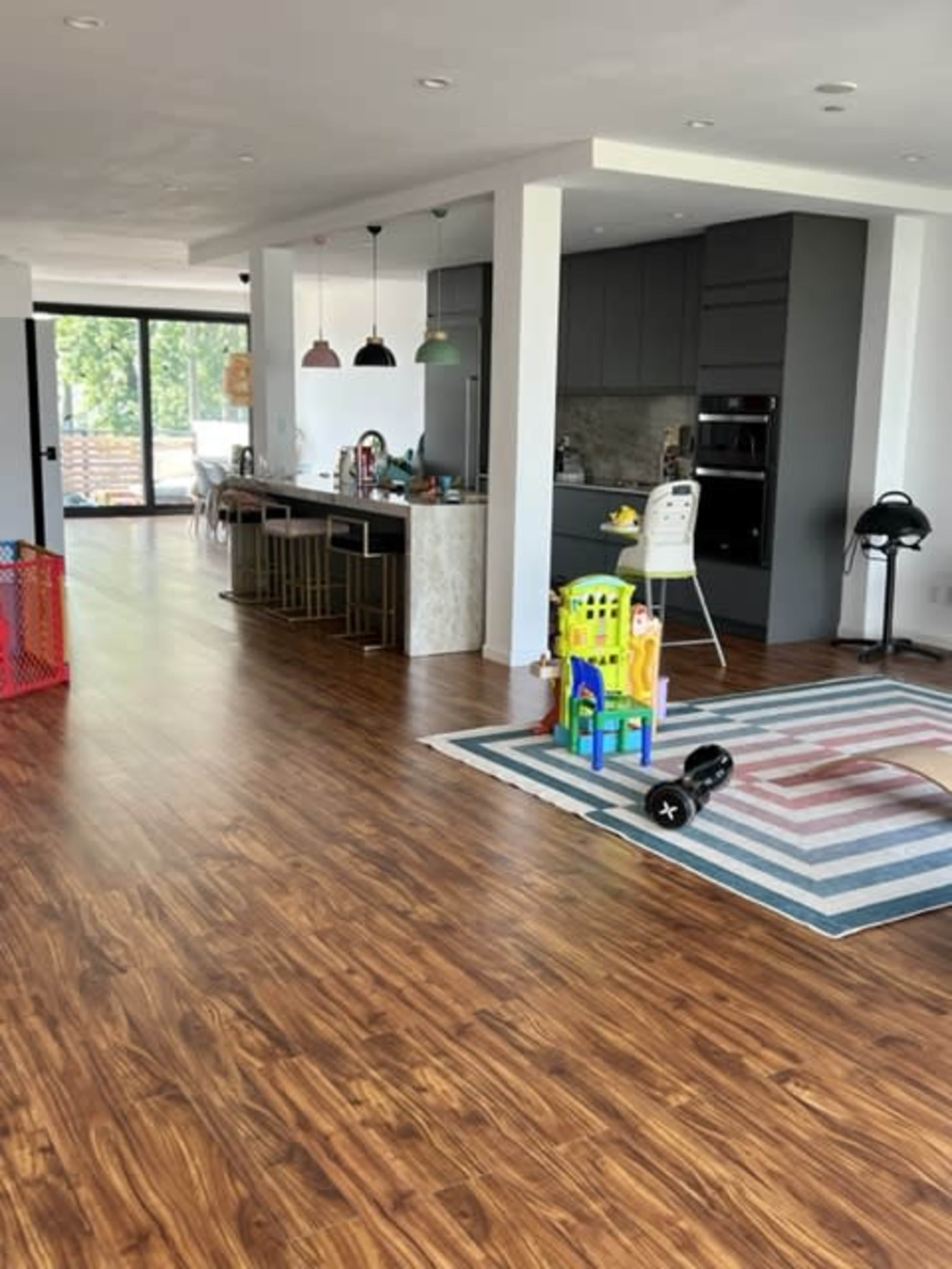 The image shows a modern open-concept living space with a kitchen on one side and a play area featuring colorful toys and a rug in the foreground.