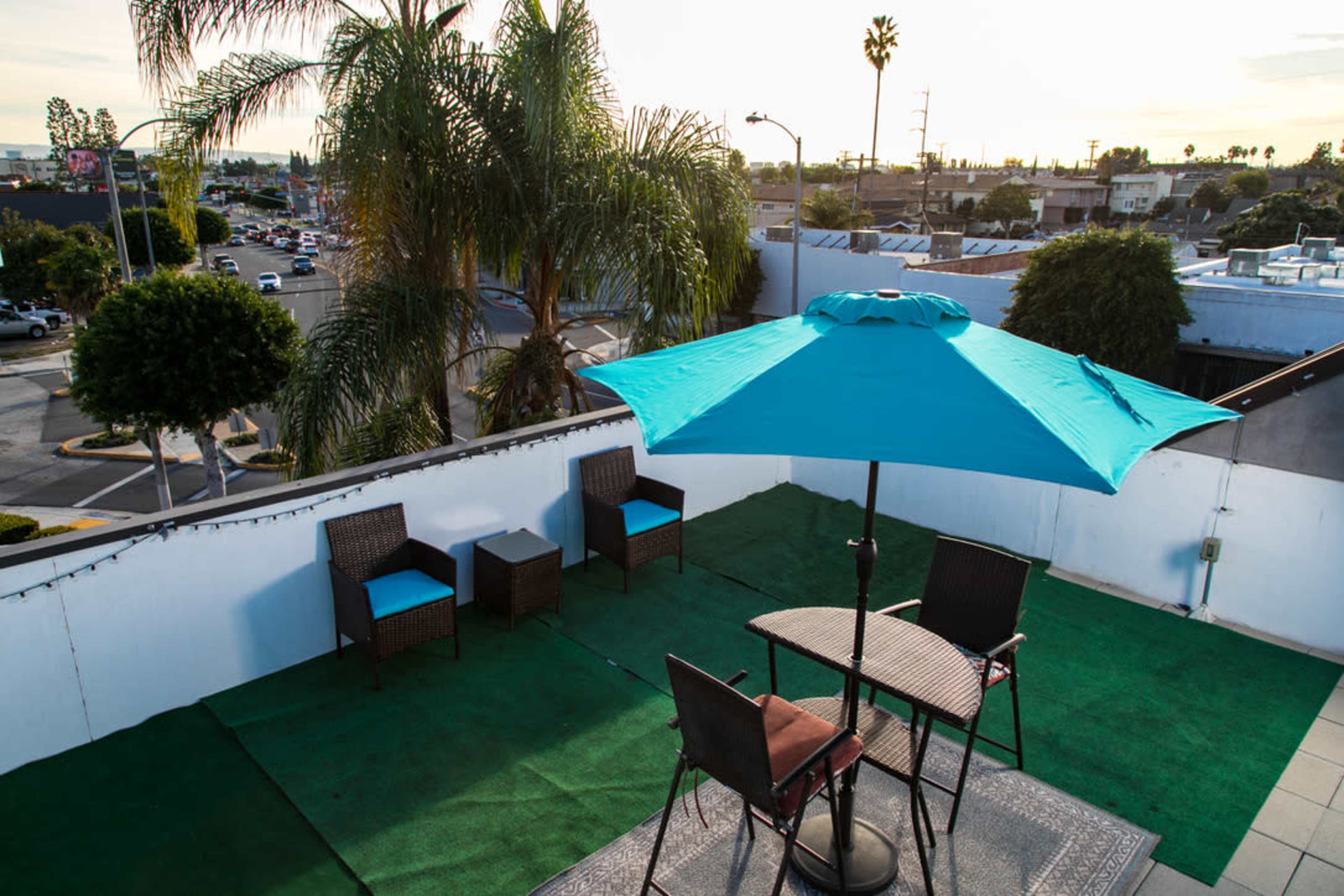 A rooftop area features a turquoise umbrella over a table and chairs, surrounded by palm trees and a view of city streets.